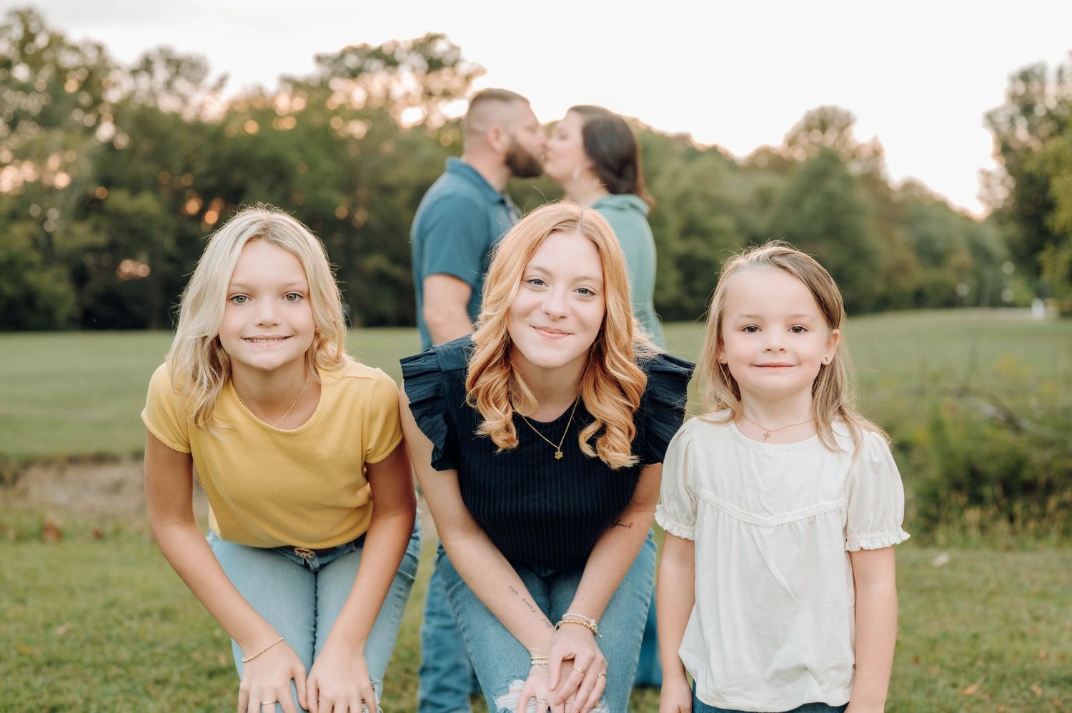 Family poses together in a grassy field during golden hour with couple kissing in background.
