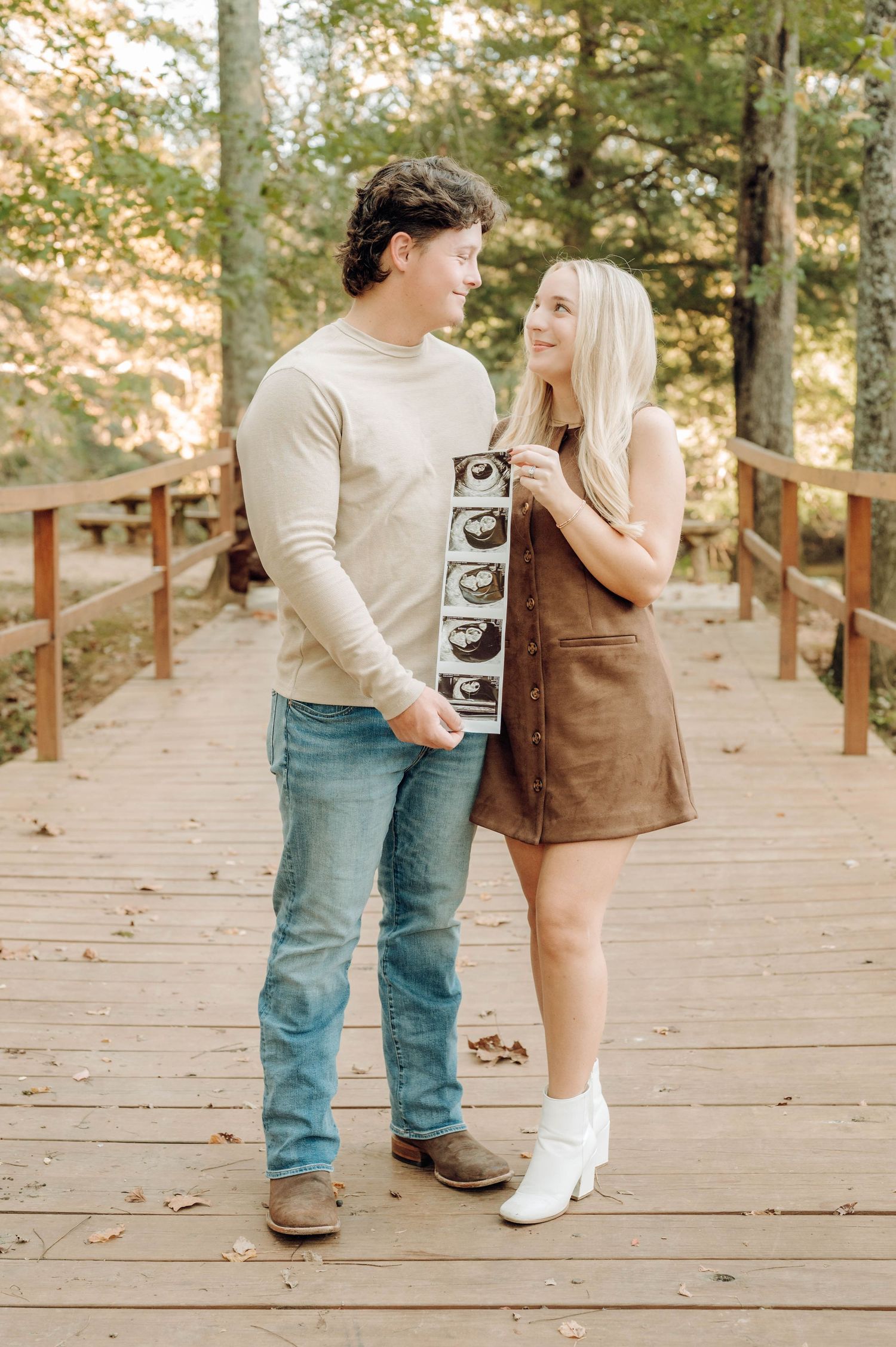 A couple stands on a wooden bridge holding ultrasound photos while sharing a romantic moment in an autumn forest setting.
