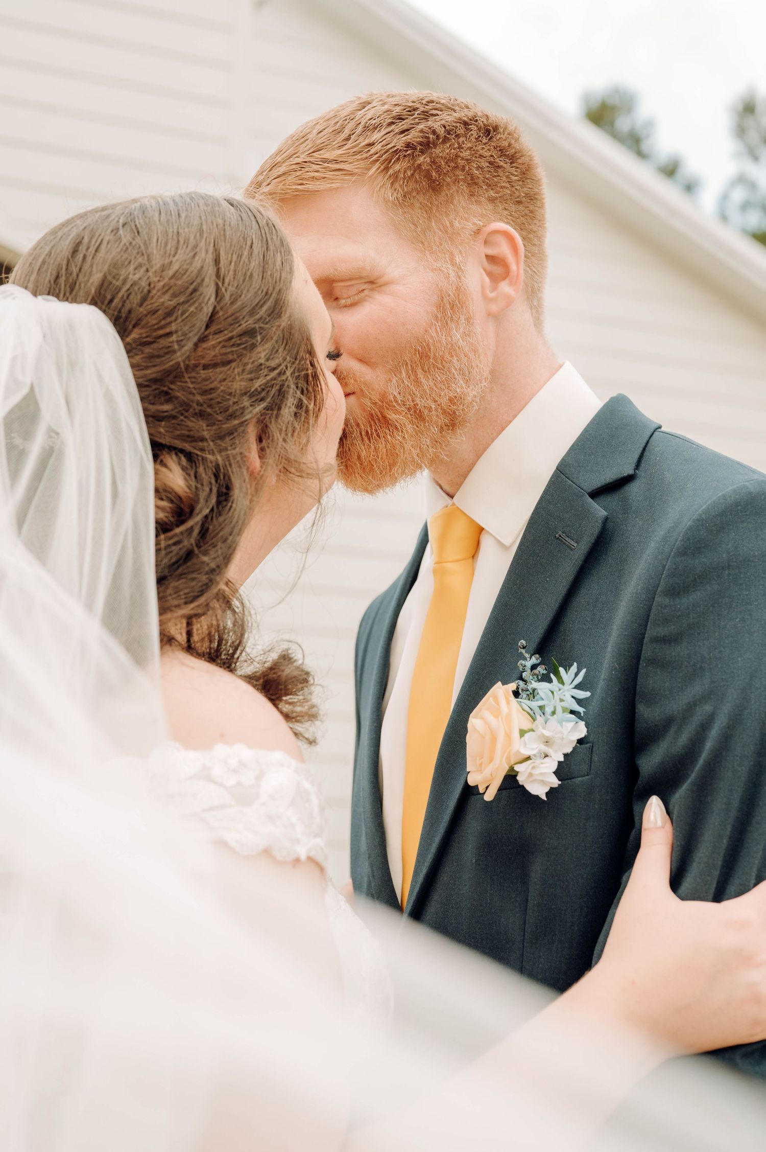 Newlywed couple sharing a tender kiss, with the bride in white veil and groom in dark suit with yellow tie and boutonniere.