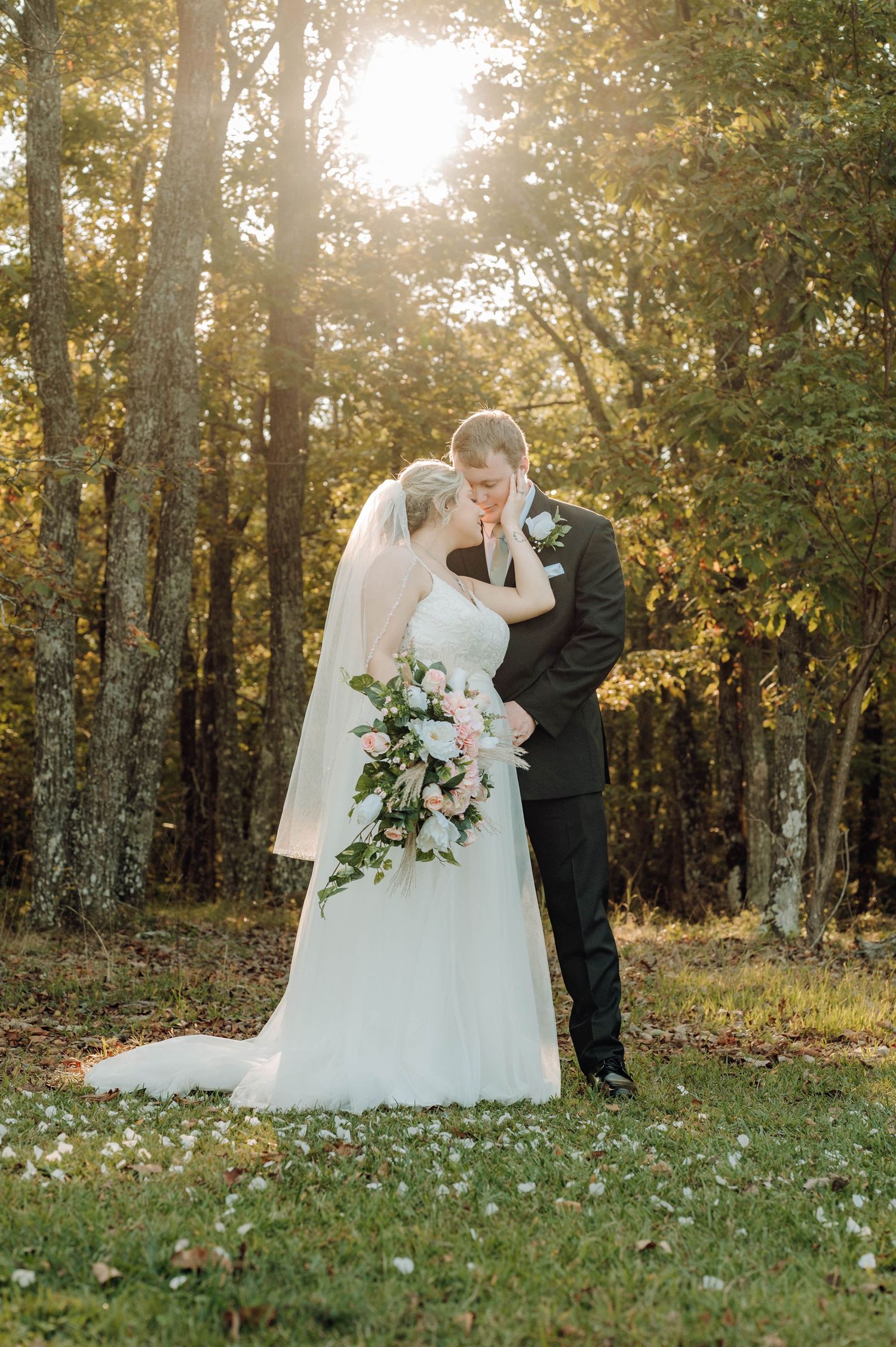A romantic wedding couple embraces in a sunlit autumn forest with scattered wildflowers on the ground.