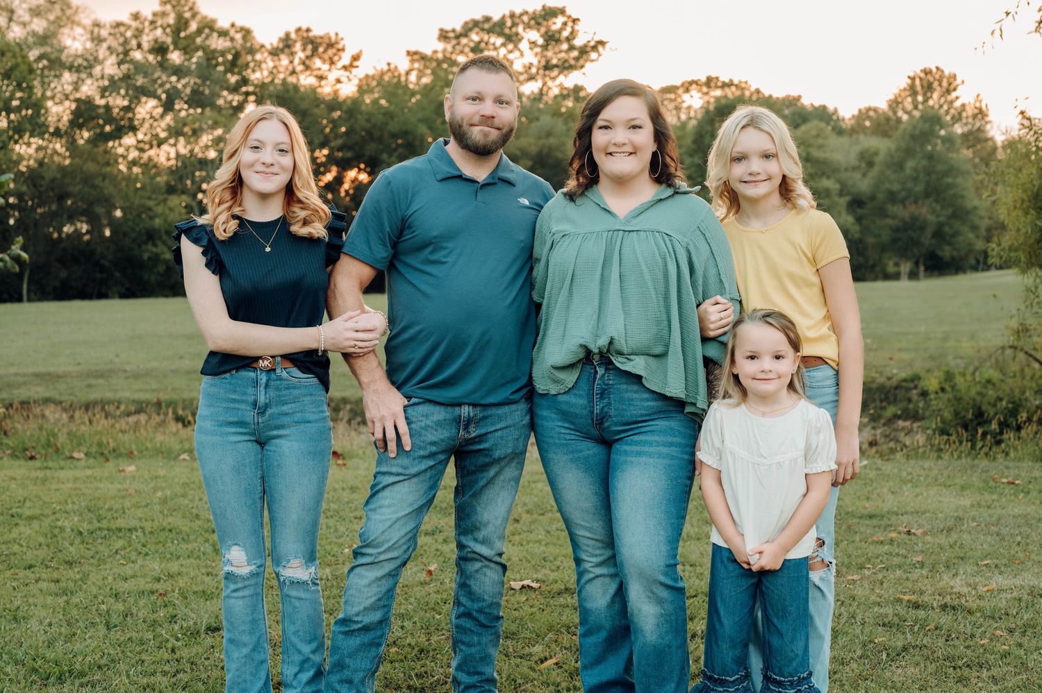A group in coordinated teal and denim outfits poses together in a sunny outdoor field at sunset.