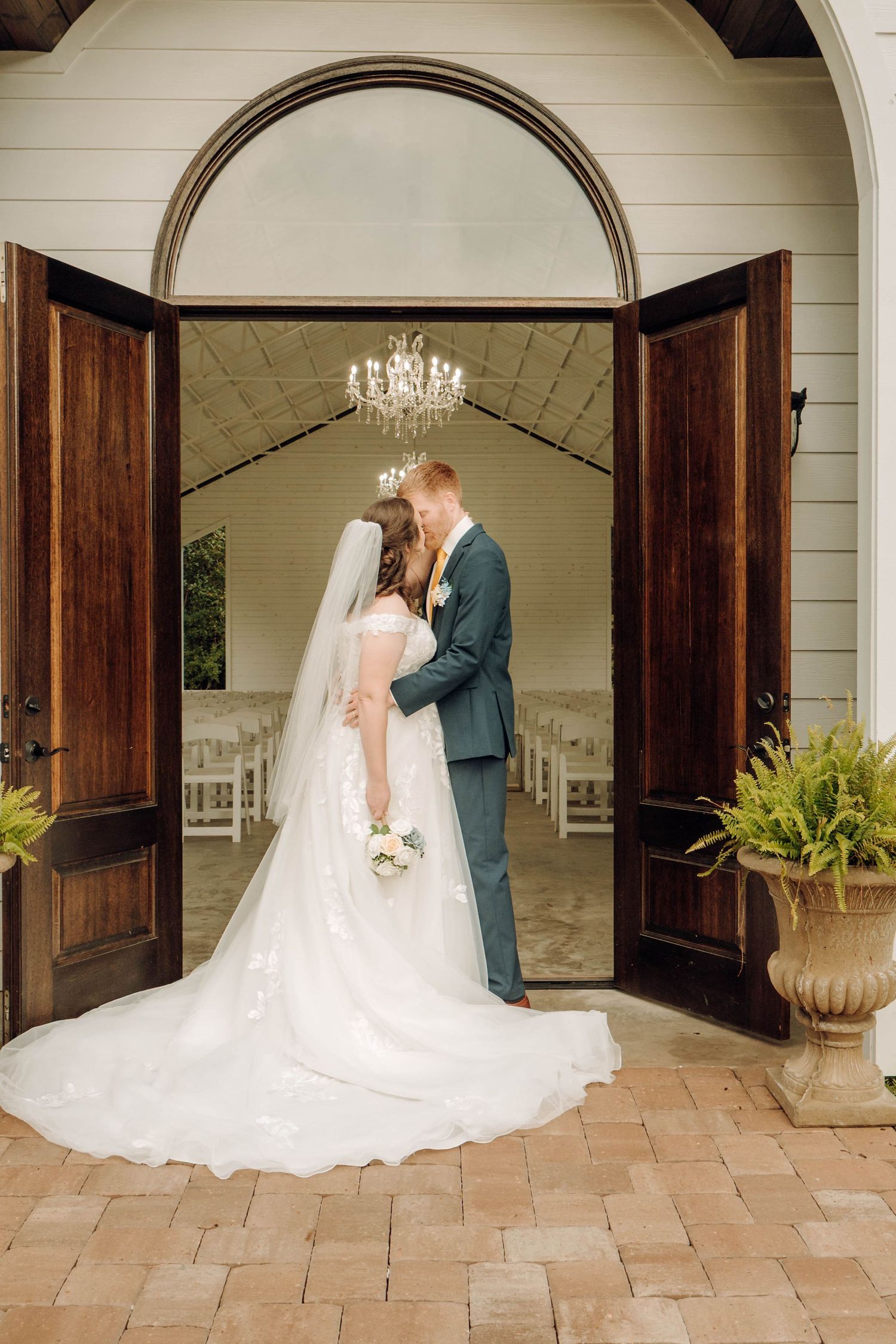 Elegant wedding couple shares a romantic moment in doorway of chapel with crystal chandelier and rustic wooden doors.