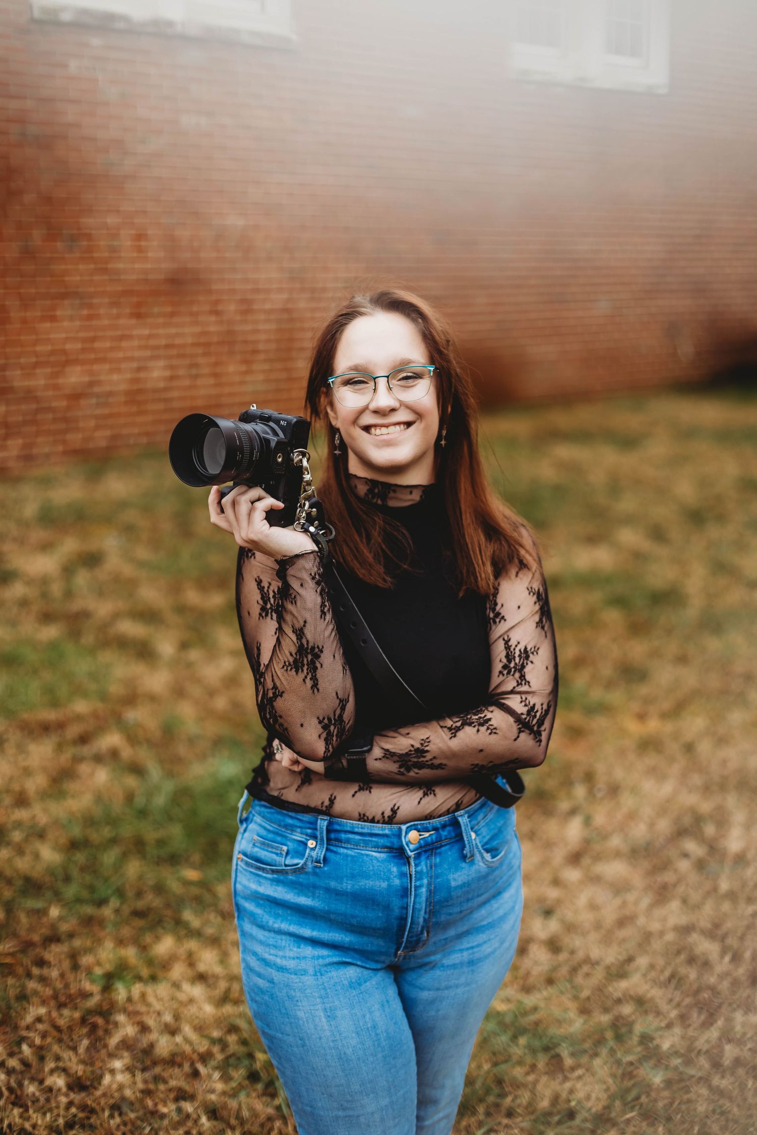 A photographer in a lace top with glasses poses with a professional camera against a brick wall.
