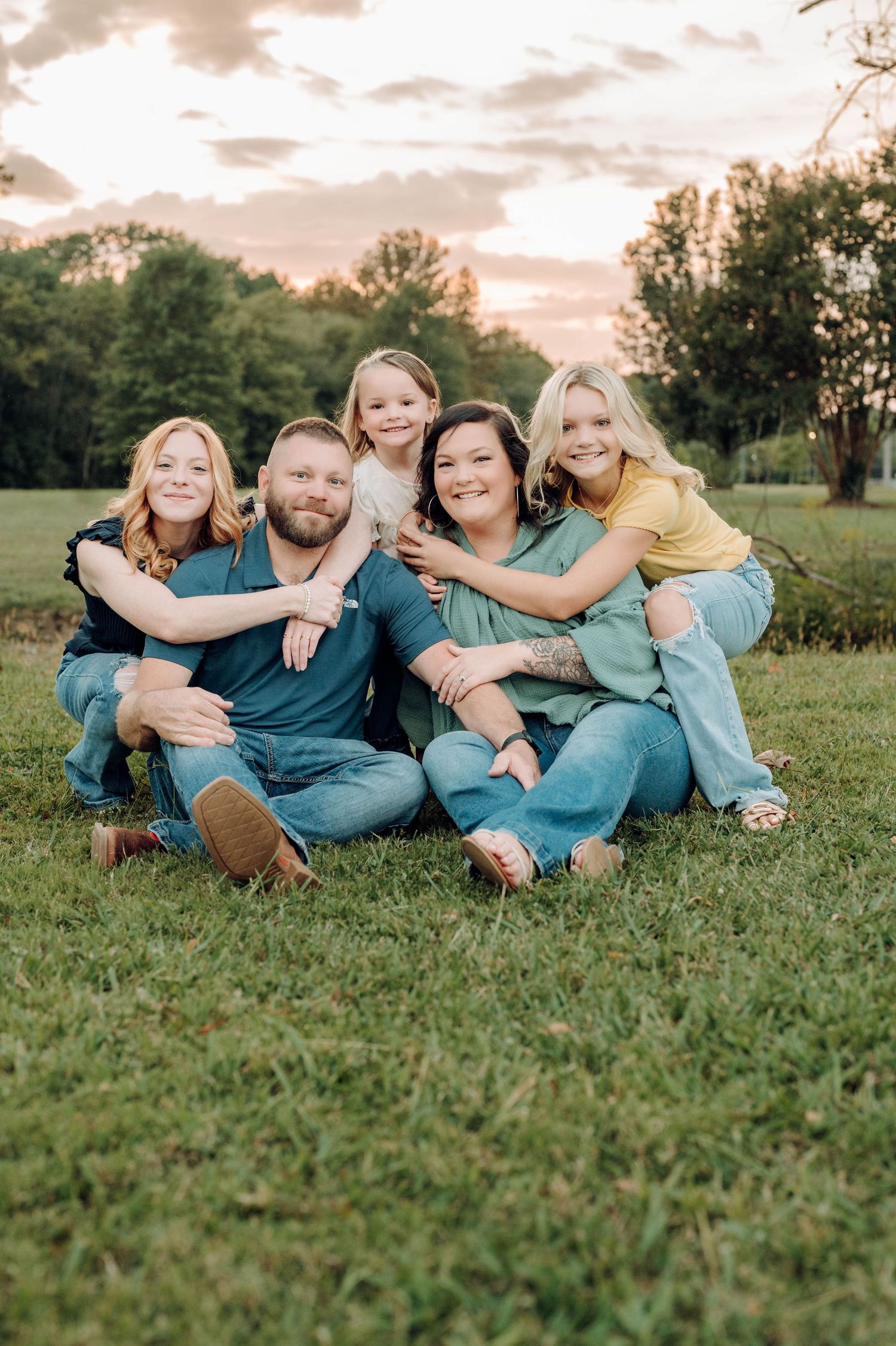 A group sits together on grass during golden hour wearing coordinating blue outfits in a casual outdoor portrait.