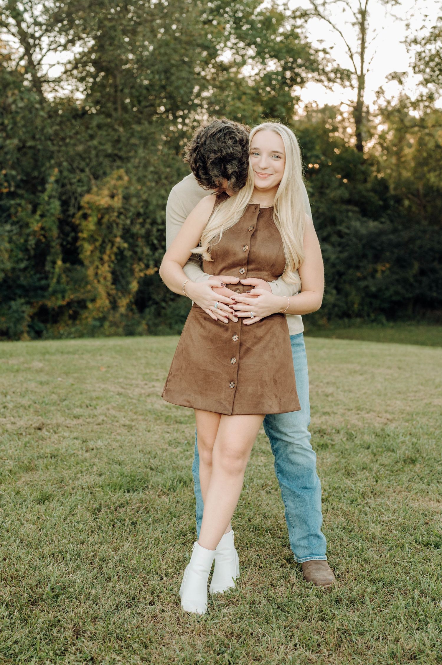 A couple embraces while holding ultrasound photos during an outdoor maternity announcement session in a grassy field.