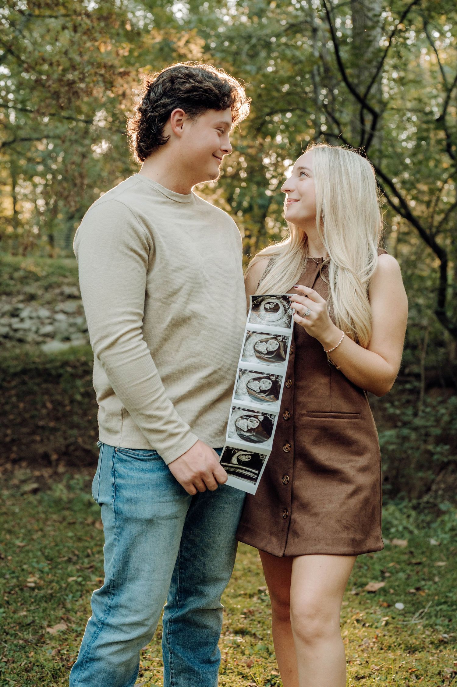 A couple poses outdoors with ultrasound photos in a romantic pregnancy announcement photoshoot in a wooded setting.