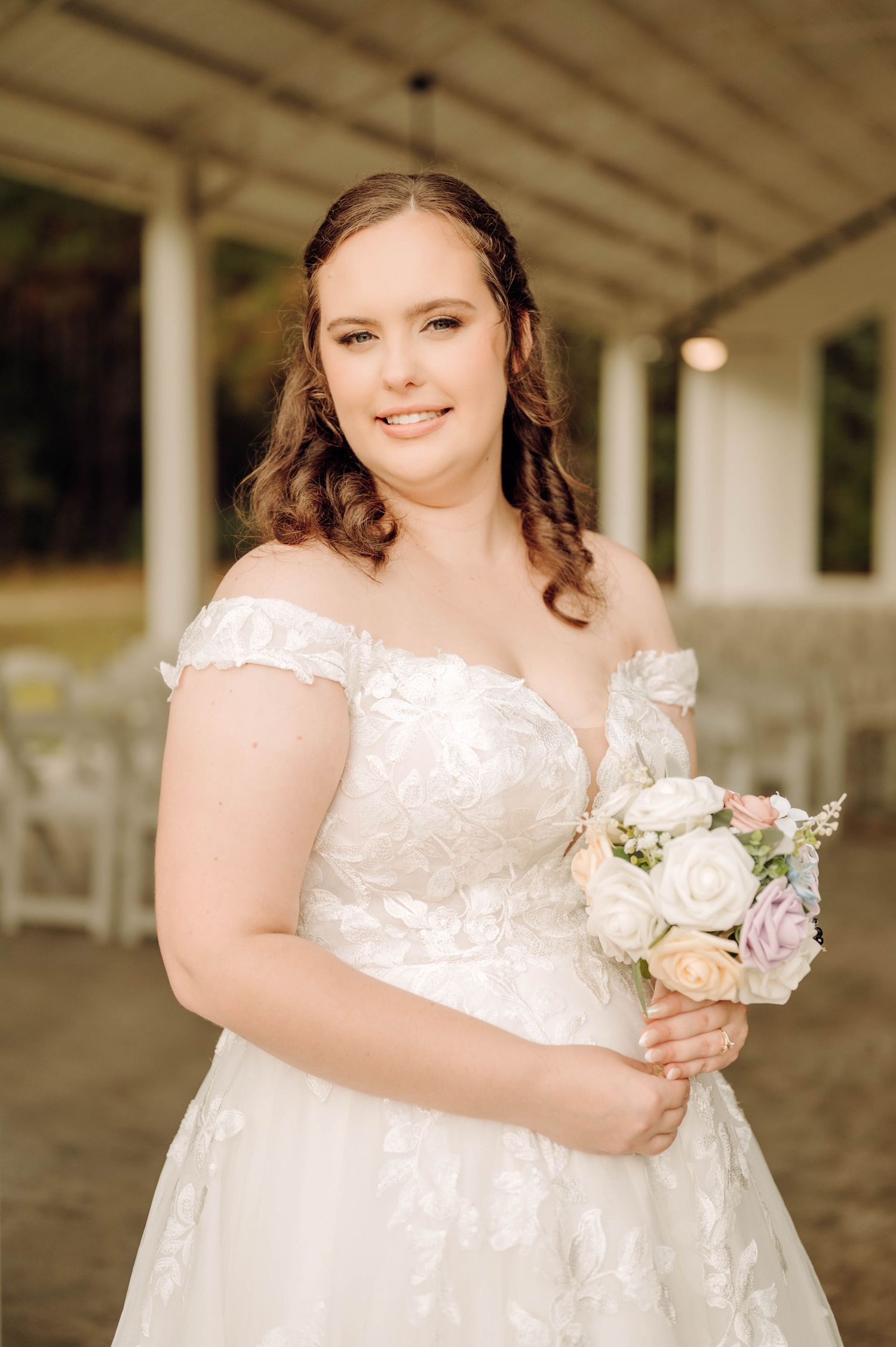 A bride in a white lace wedding dress holds a delicate bouquet of light-colored flowers in an outdoor covered venue.