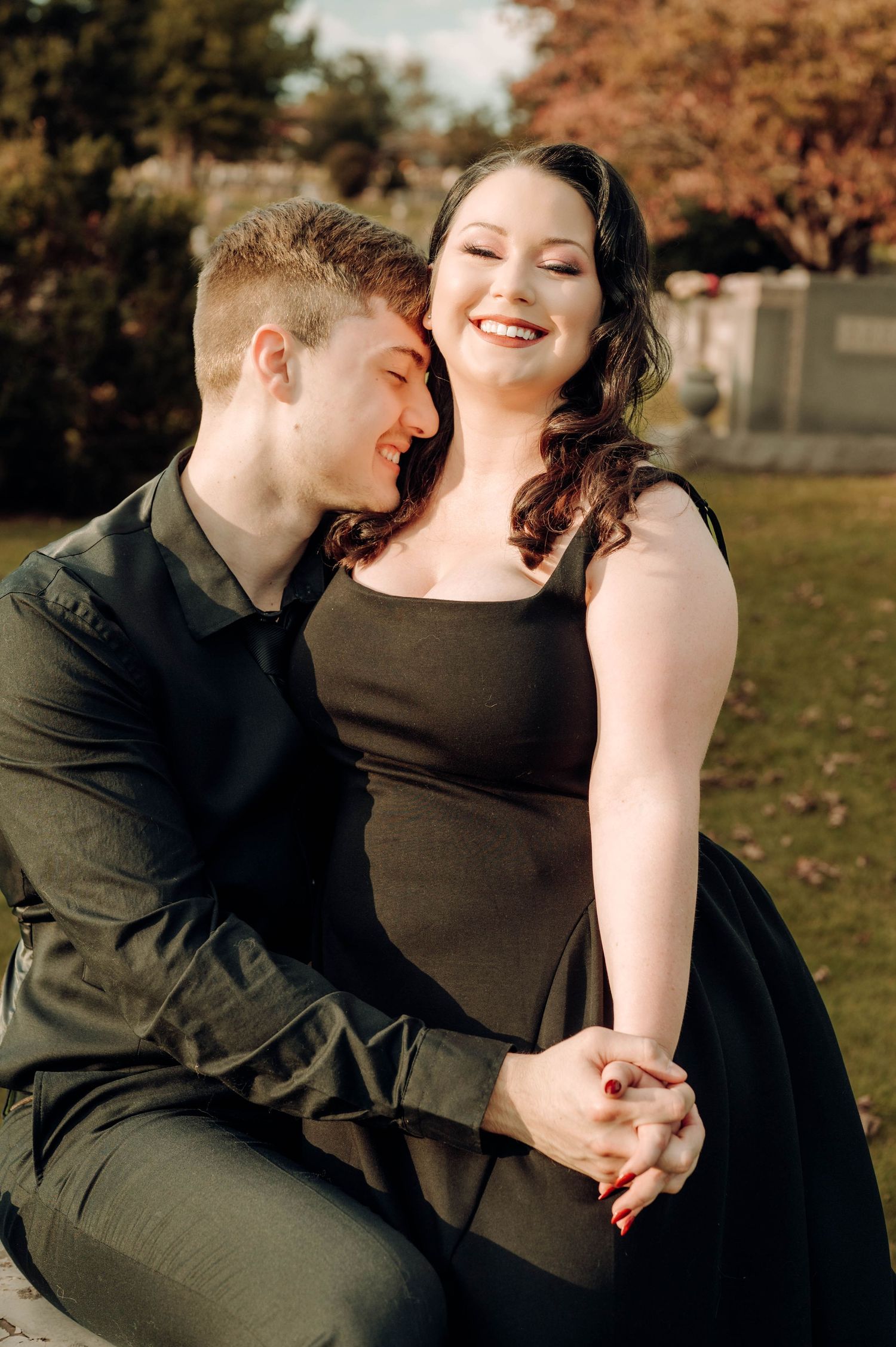 A couple in black attire shares intimate moments on autumn grass during an outdoor photoshoot.