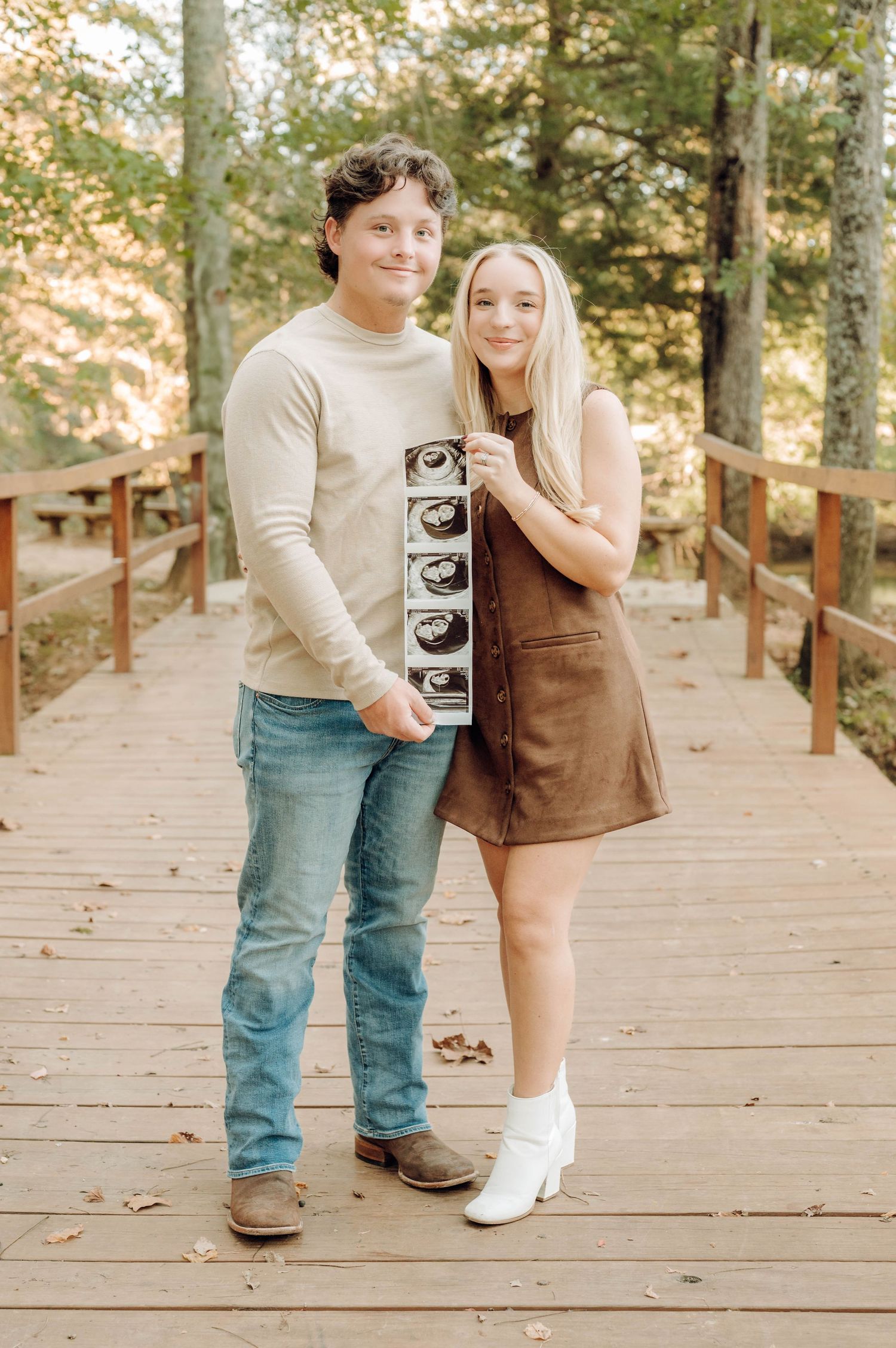 A couple stands on a wooden bridge in autumn holding ultrasound photos while sharing a joyful moment together.