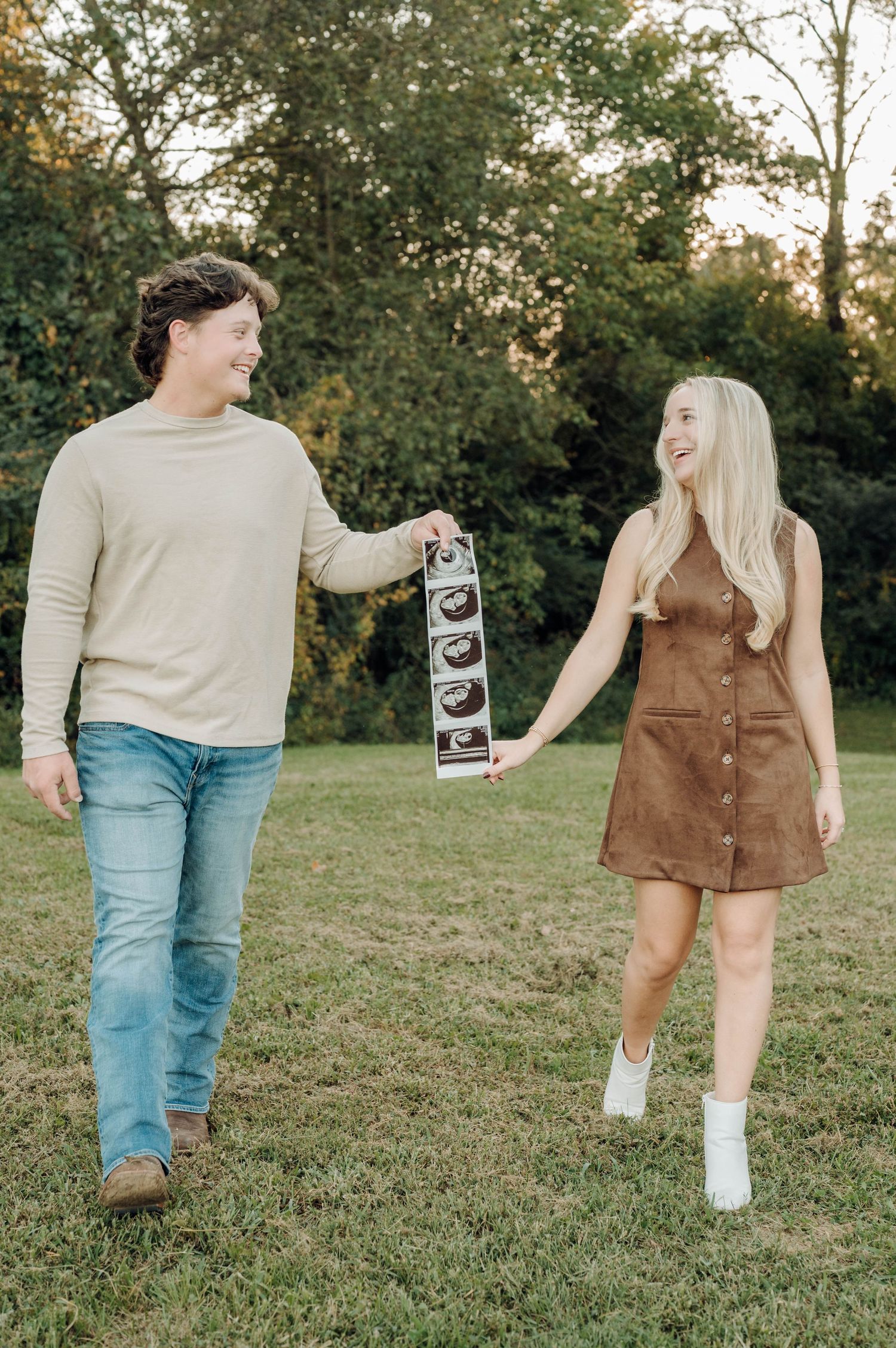 A couple stands in a grassy field holding ultrasound photos, wearing casual fall attire during a pregnancy announcement photoshoot.