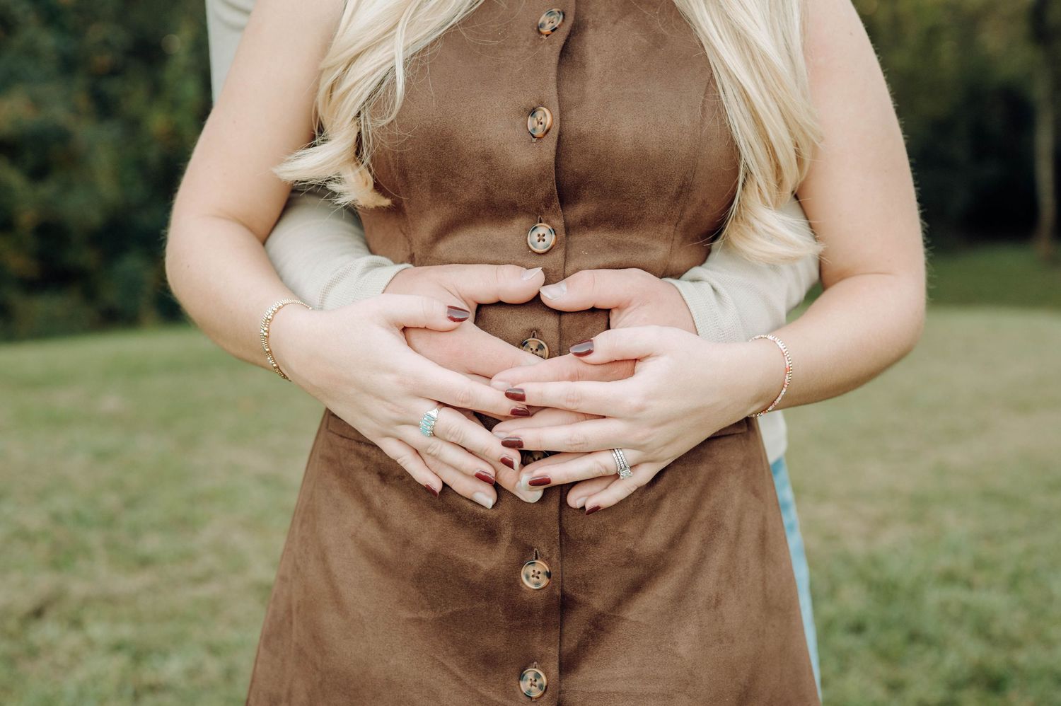 Close-up view of a brown button-up dress with hands forming a heart shape against a grassy outdoor background.