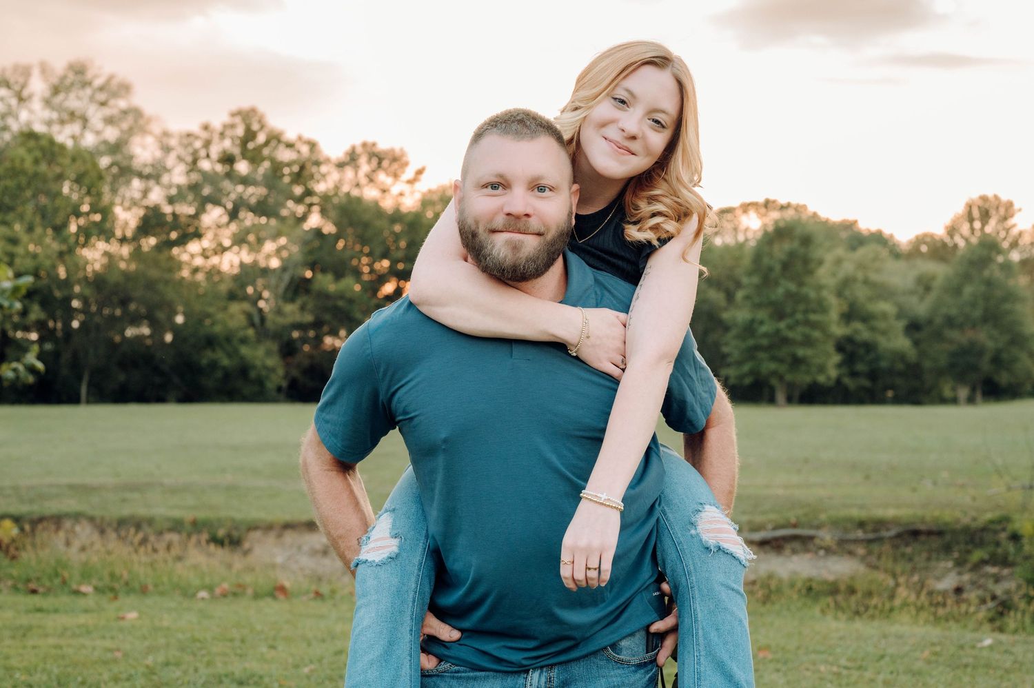 A couple in teal outfits poses together in a sunny field during golden hour for engagement photos.