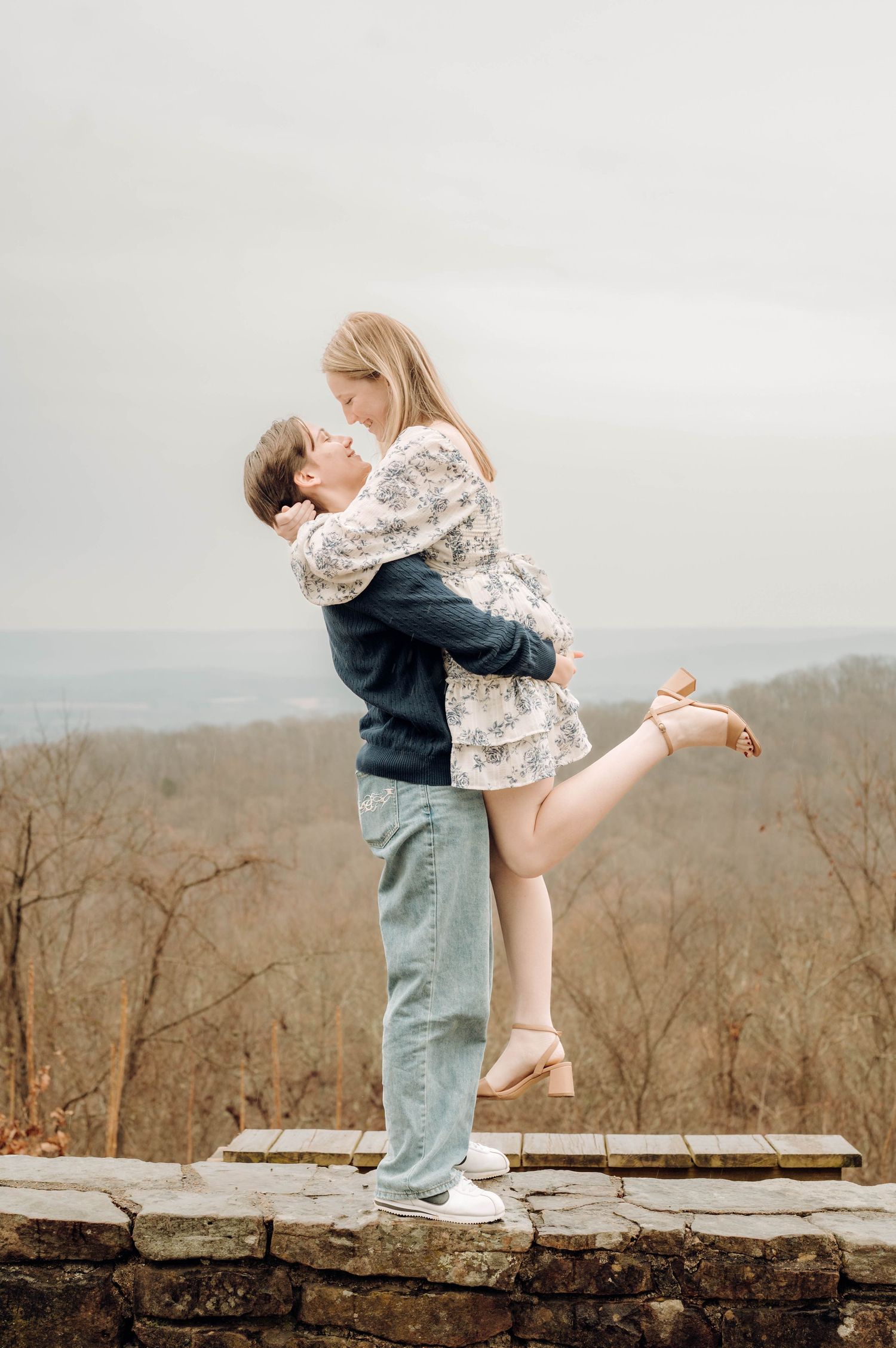 A romantic couple embraces on a stone wall overlooking misty mountains, with the woman lifting one leg in a playful pose.