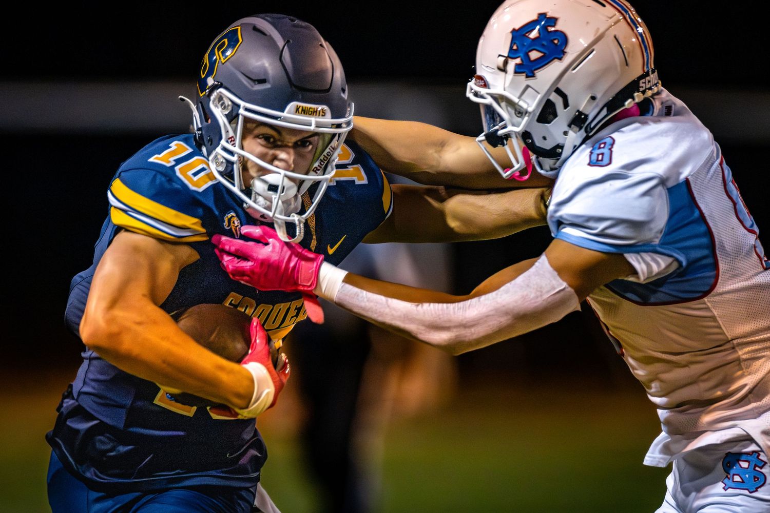 Two football players collide during a tackle attempt in a nighttime game under stadium lights.