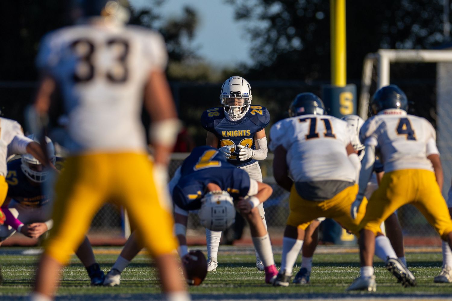High school football game shows an offensive play setup with players in white and dark uniforms at sunset.