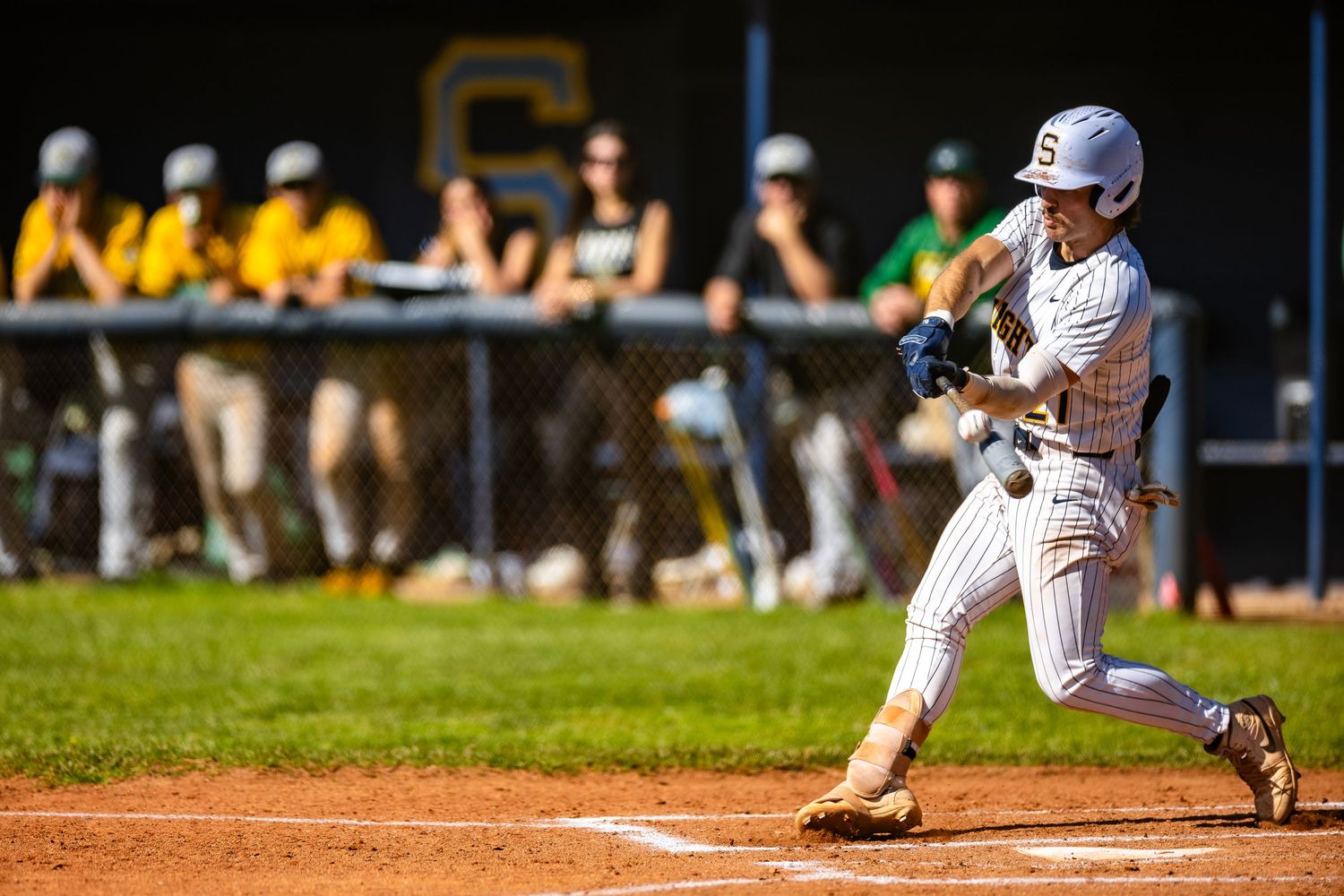 Baseball player in white pinstriped uniform swings the bat during a game with spectators watching from the dugout area.