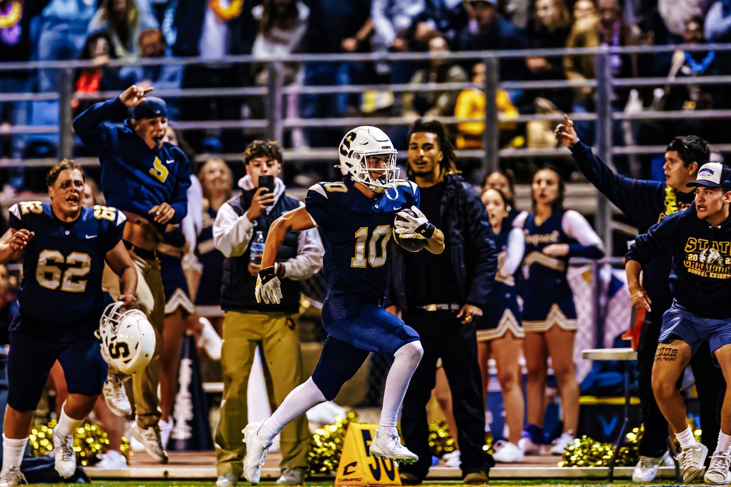 Football players celebrate and cheer from the sidelines during an intense night game at a stadium.