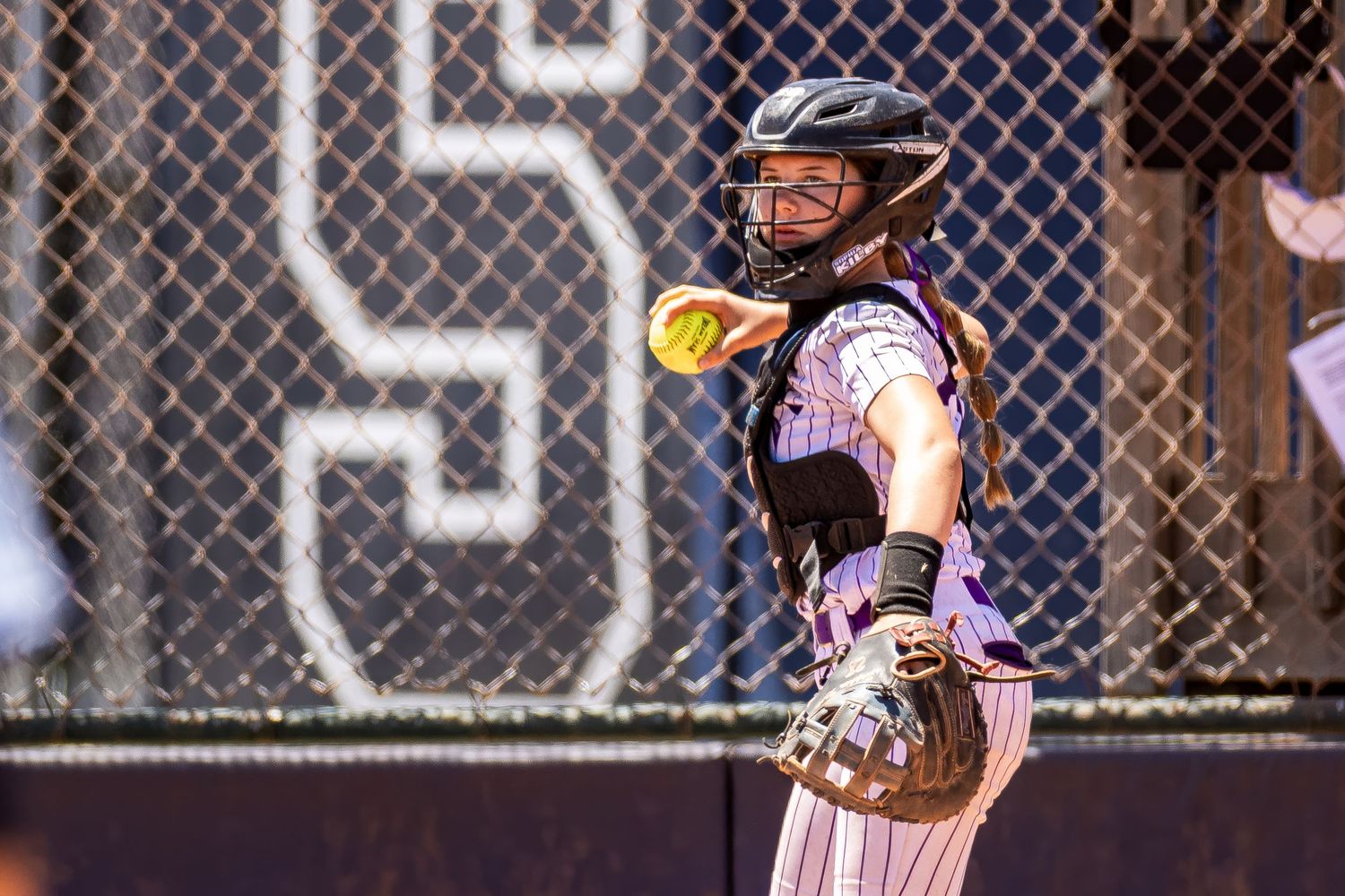A softball catcher in protective gear looks on during a game behind home plate.