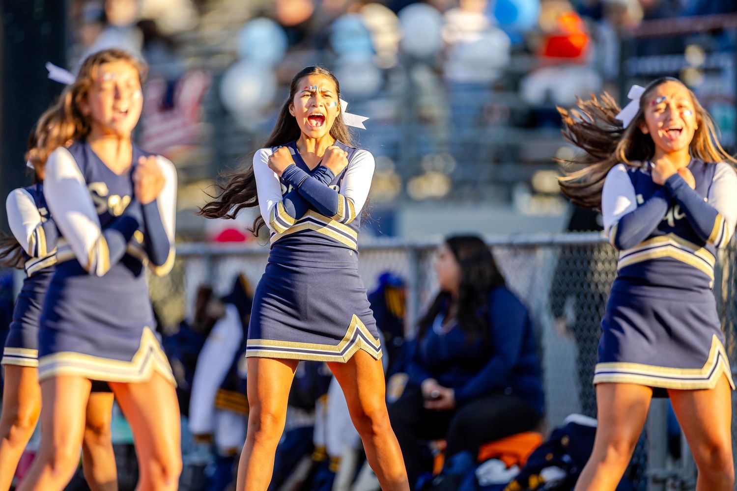 A group of cheerleaders in navy and white uniforms perform synchronized dance moves during an outdoor sporting event.