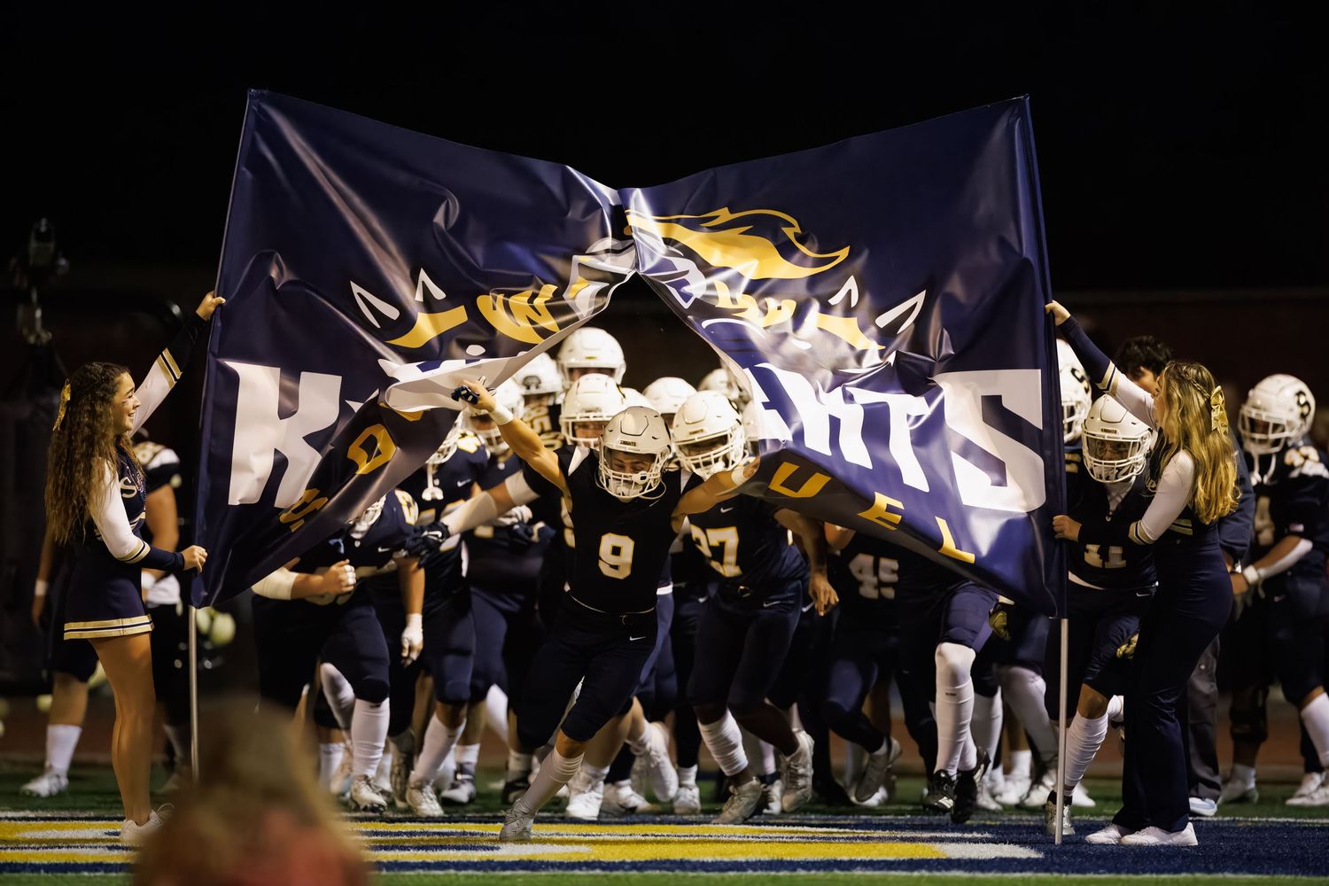 High school football team charges through banner during nighttime game celebration.