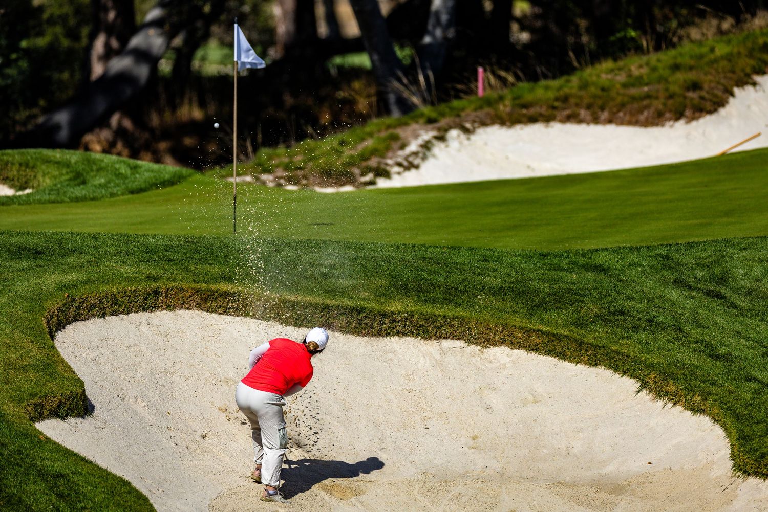 Golfer on a scenic course prepares to hit from a white sand bunker near a well-manicured green.