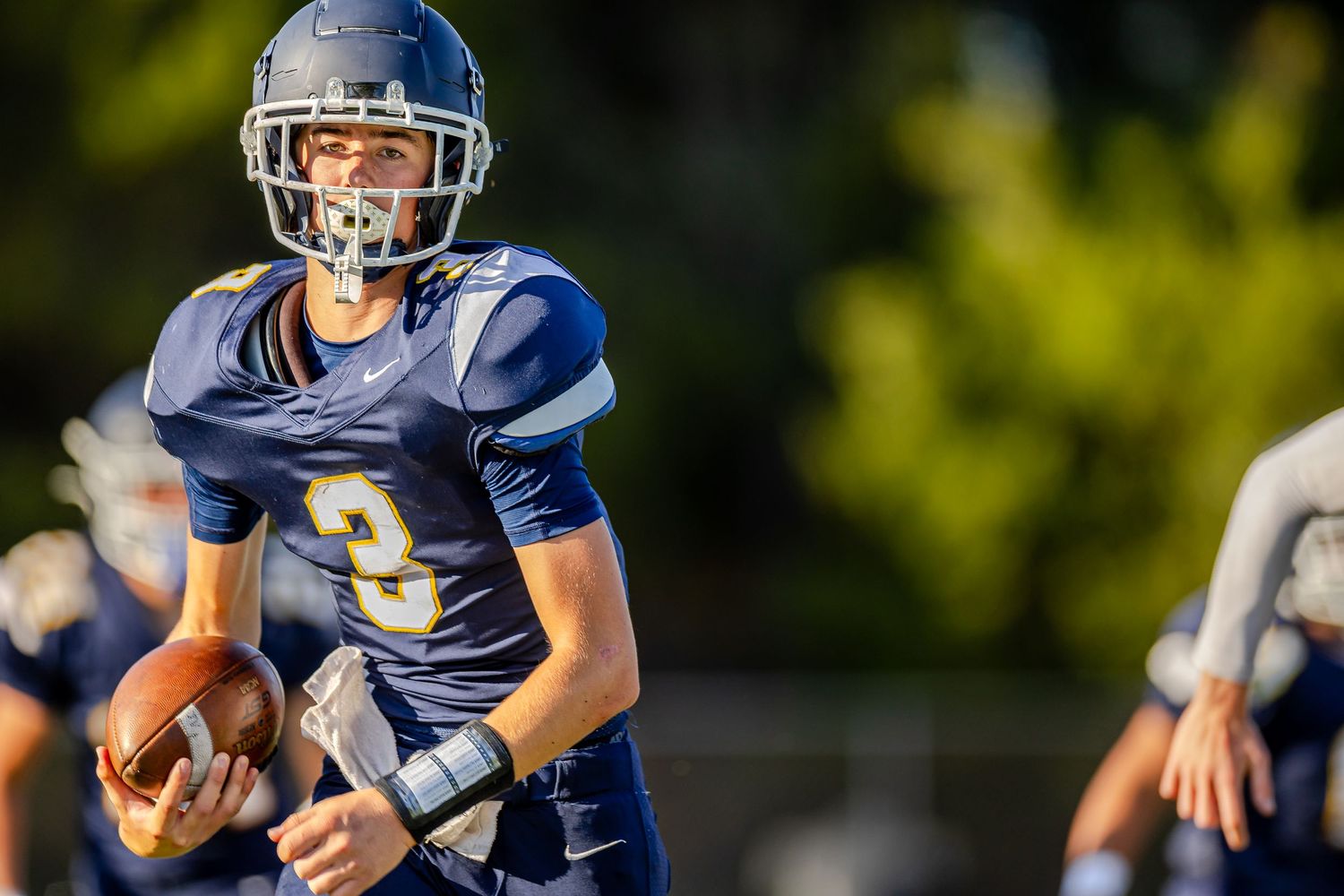 A football player in blue uniform runs with the ball during an outdoor game with trees in the background.