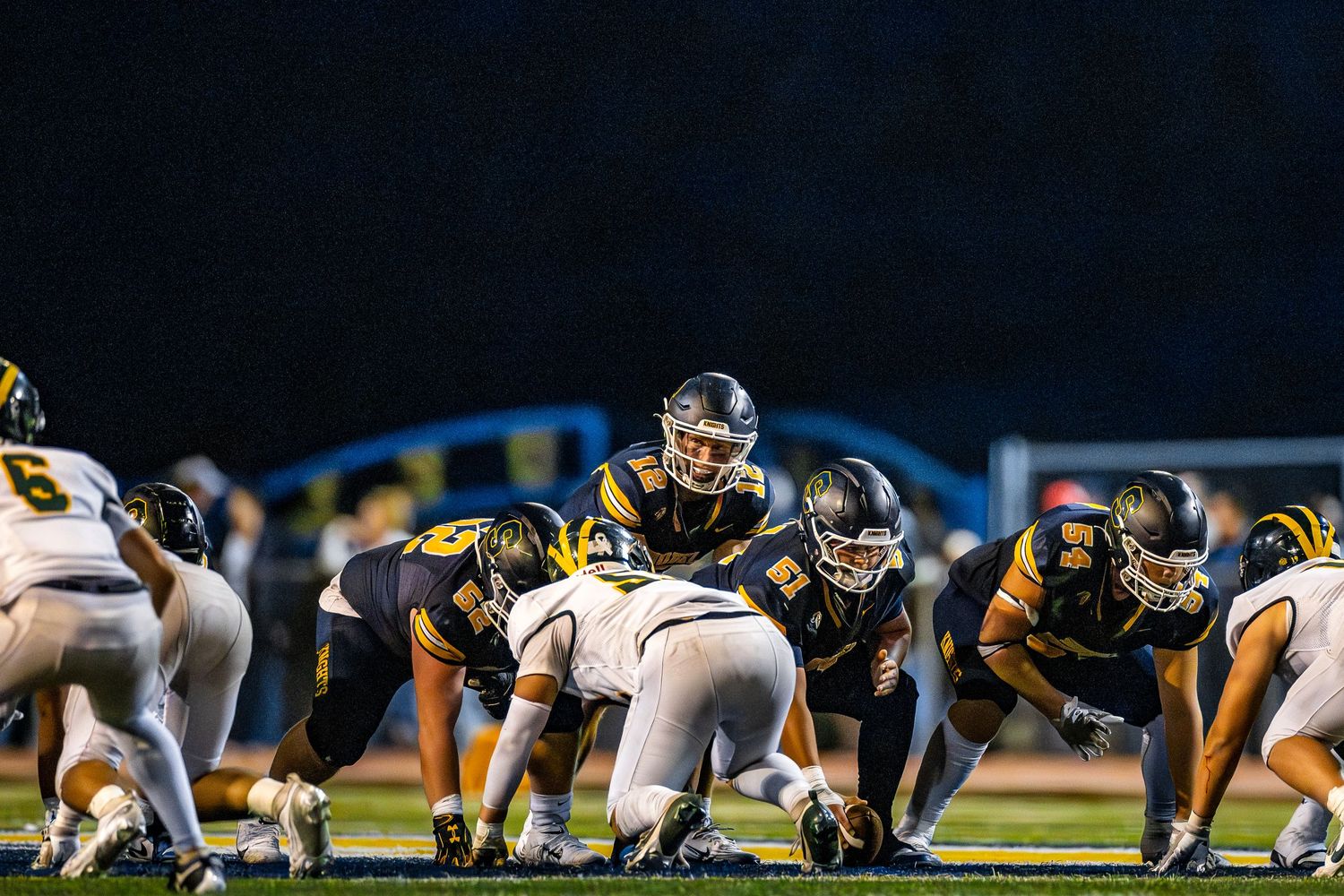 Football teams line up in formation at the line of scrimmage during a night game under stadium lights.