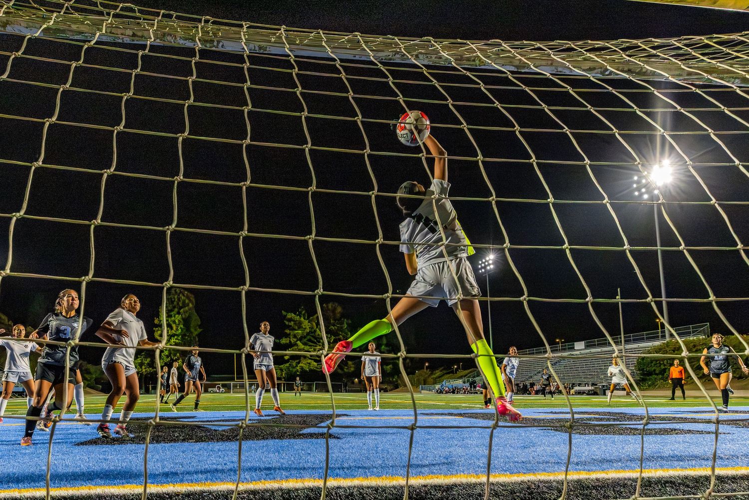 Soccer goalkeeper stretches to make a save at the goal net during a nighttime soccer match.