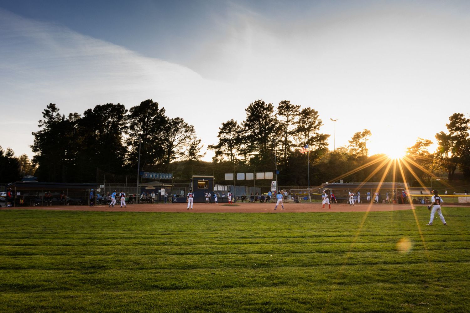 Baseball field at sunset with sun rays streaming through trees and spectators in background.