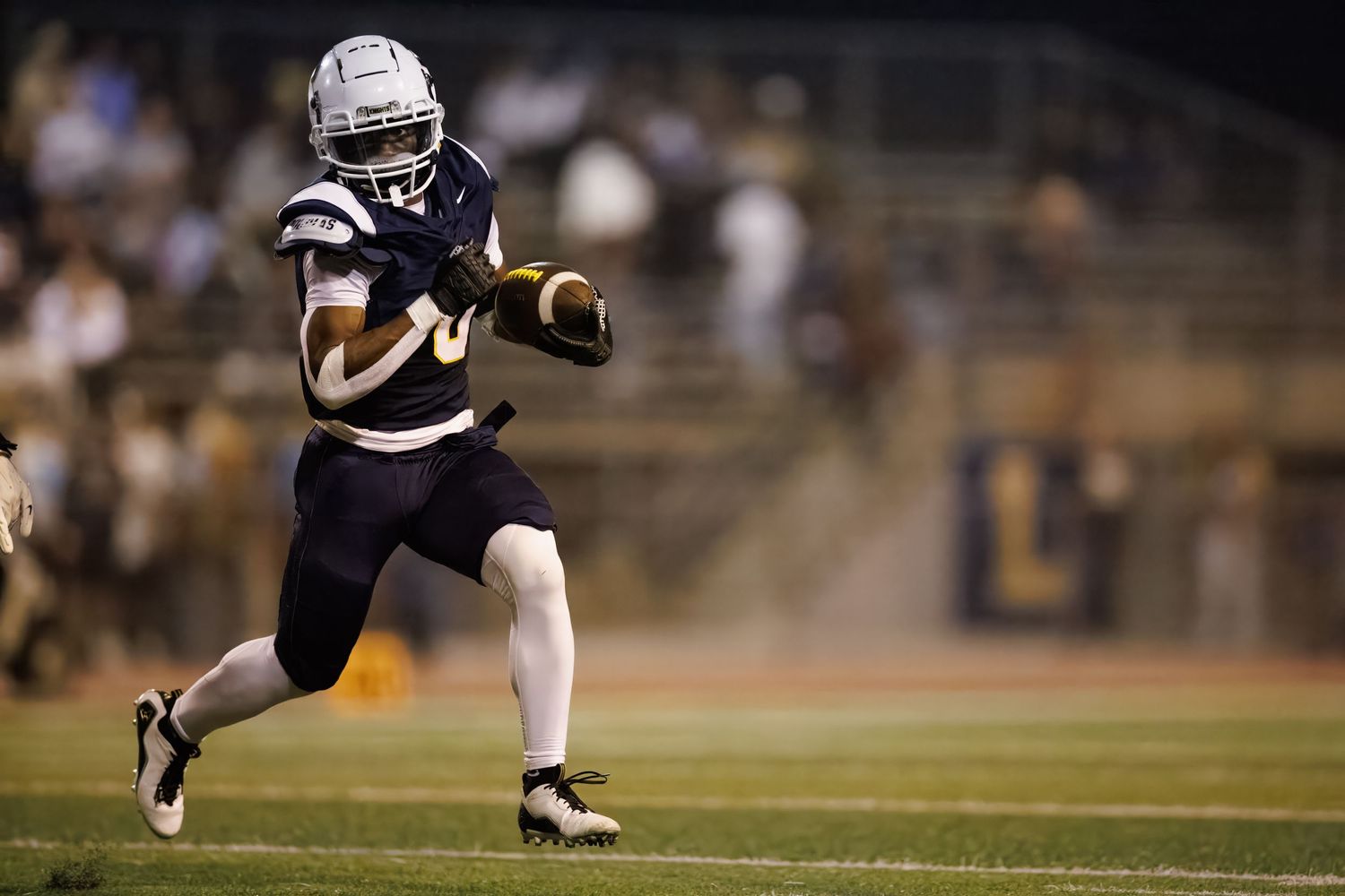 Football player in dark uniform runs with ball during nighttime game on illuminated field.