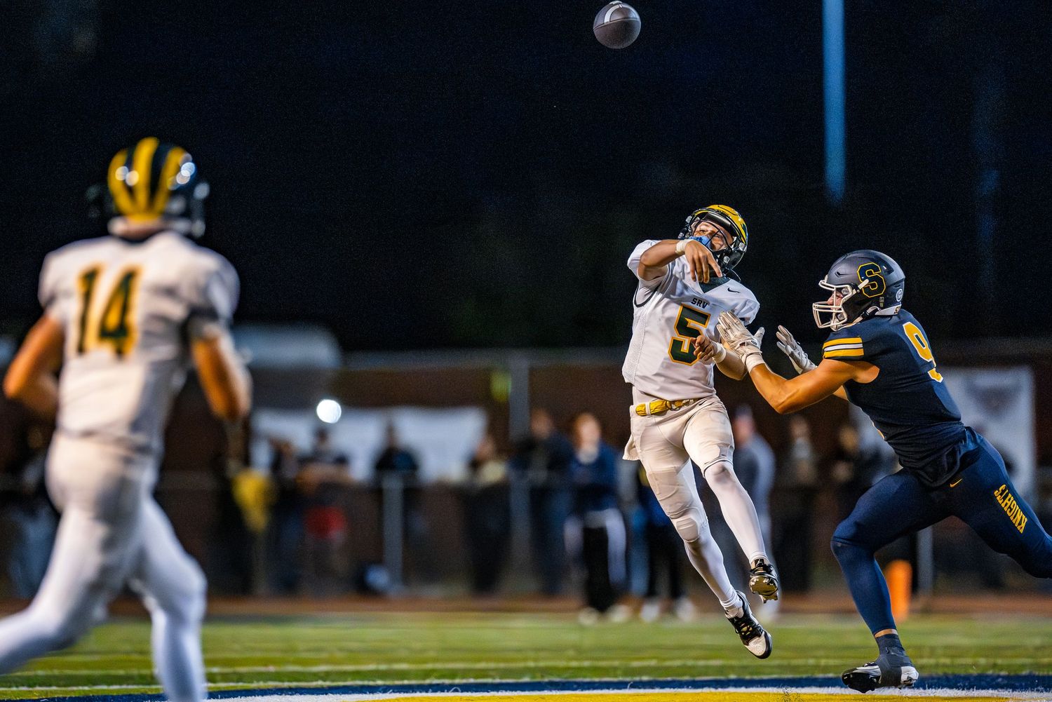 Multiple football players in yellow and blue uniforms compete on a dark field during a nighttime game.
