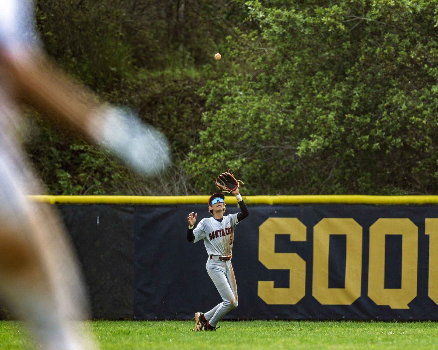 An outfielder makes a catch near the wall at a baseball stadium with 'SOON' visible in large letters.