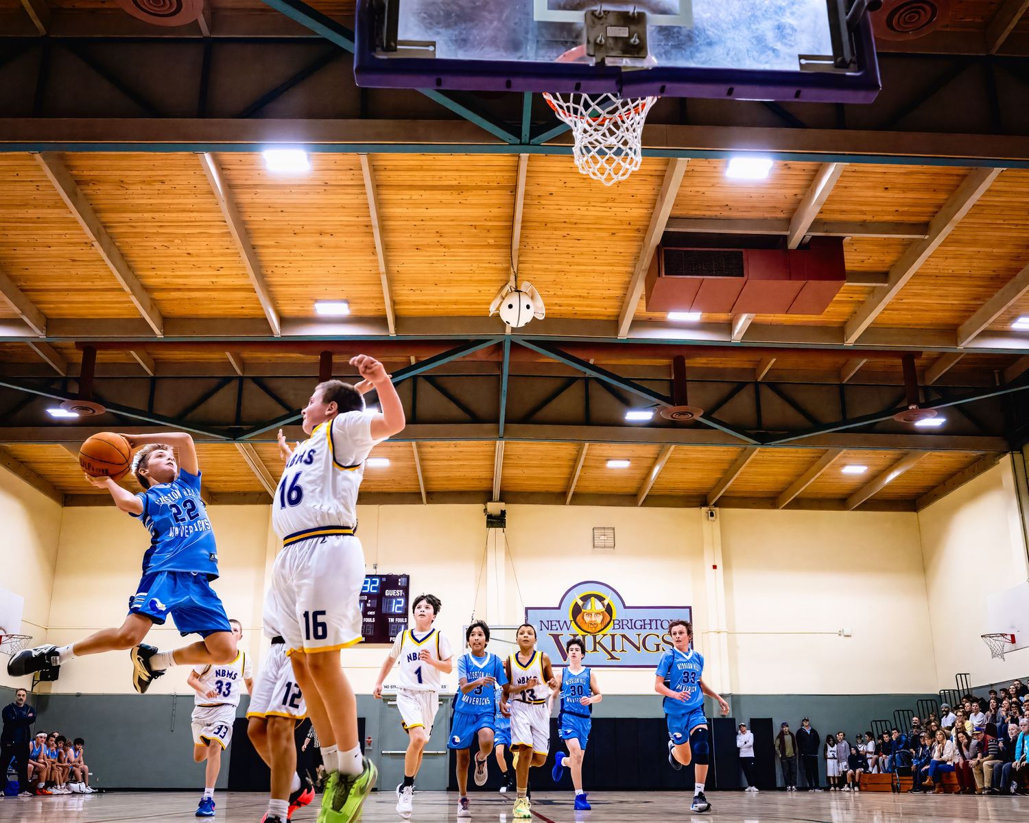Two basketball teams compete in an indoor gym with wooden ceiling beams as a player attempts a layup shot.