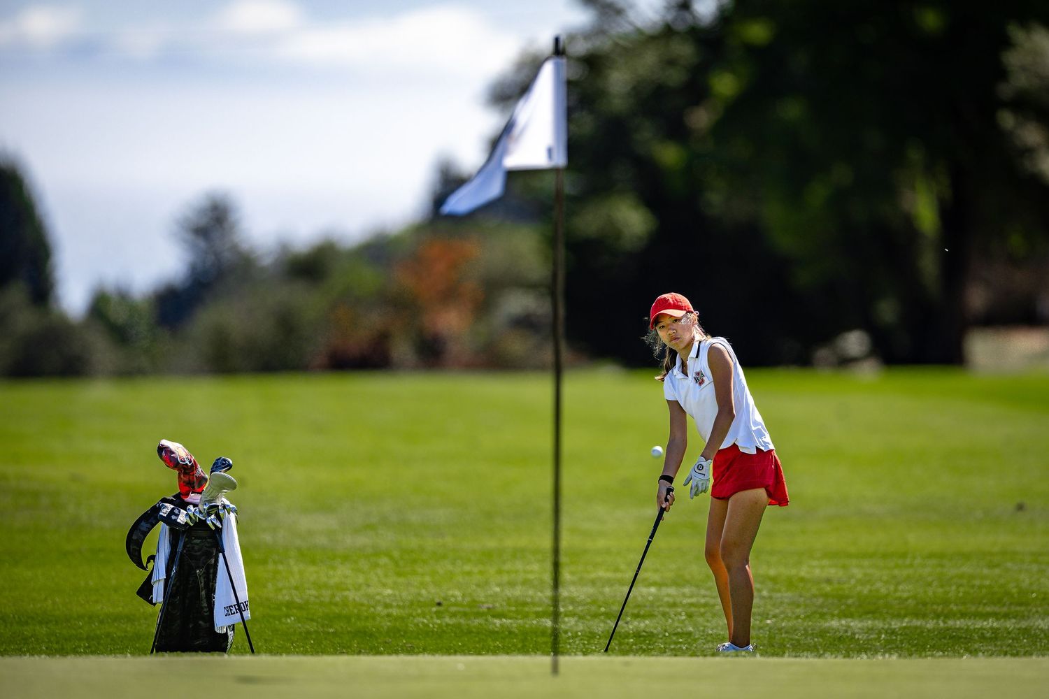 A golfer lines up a shot next to a flag on a sunny golf course with golf bag visible in background.