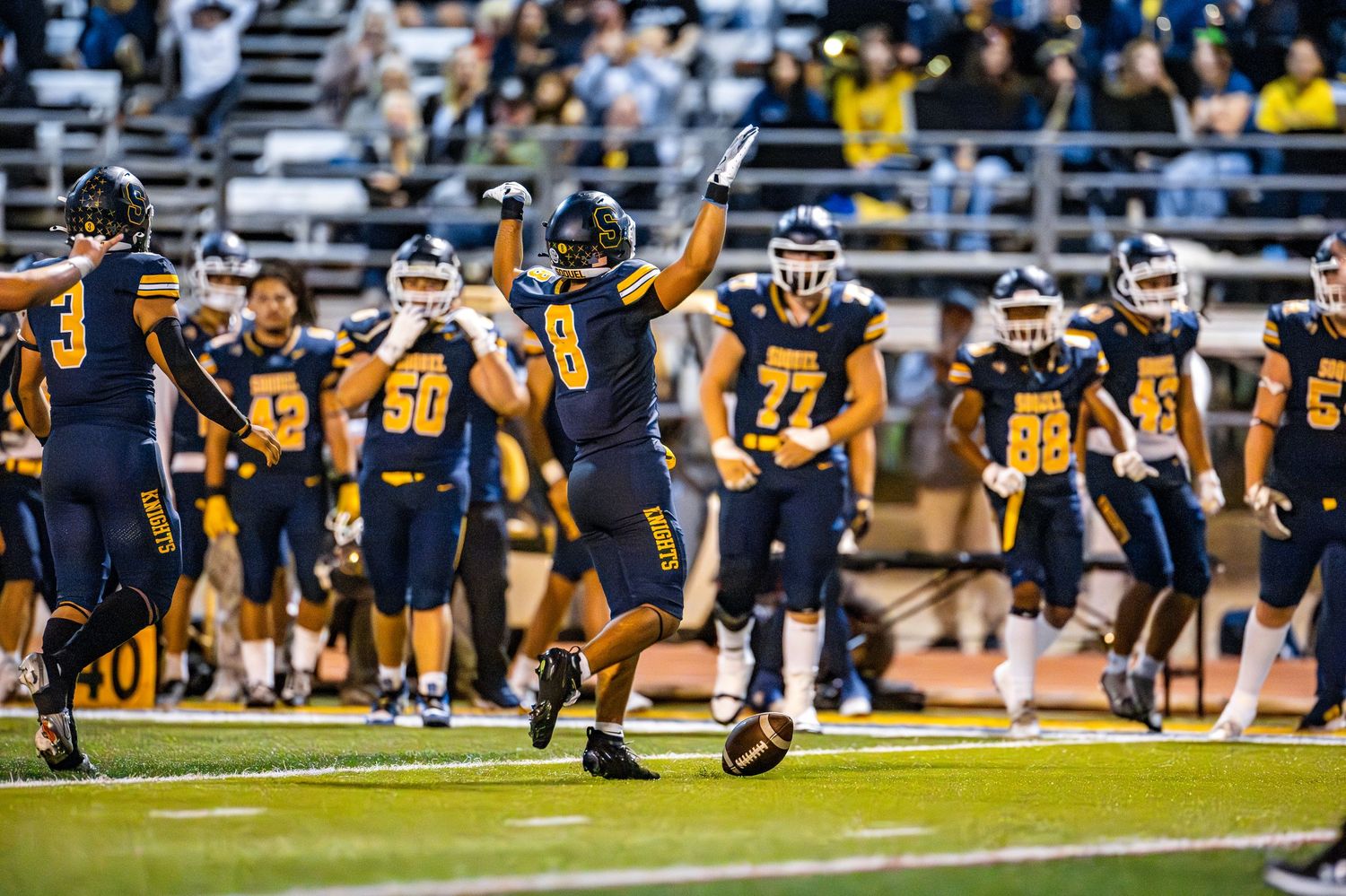 Football players on the sidelines watch intently during a game in blue and yellow uniforms.