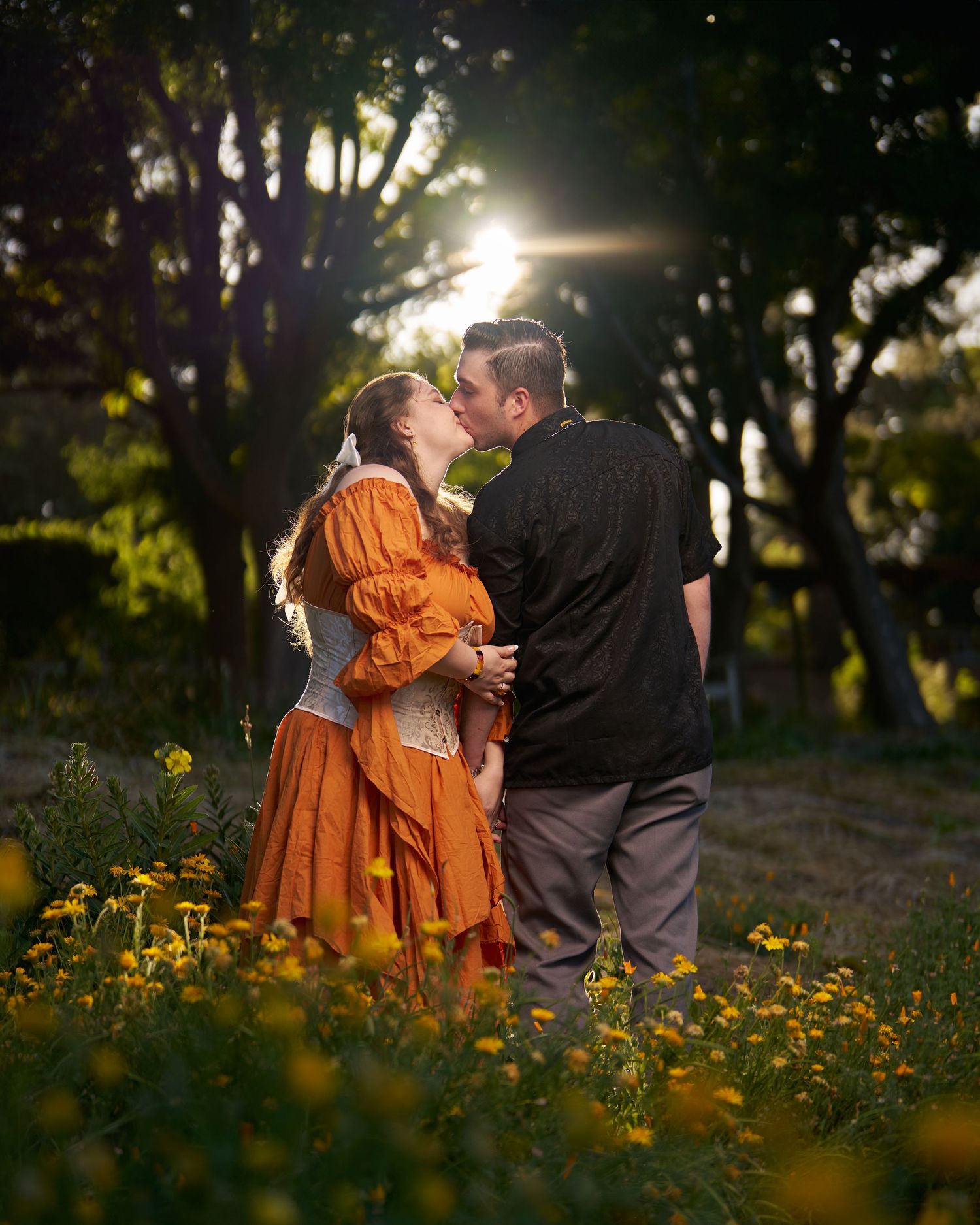 A newly engaged couple kissing in a flower field with the sun behind them.