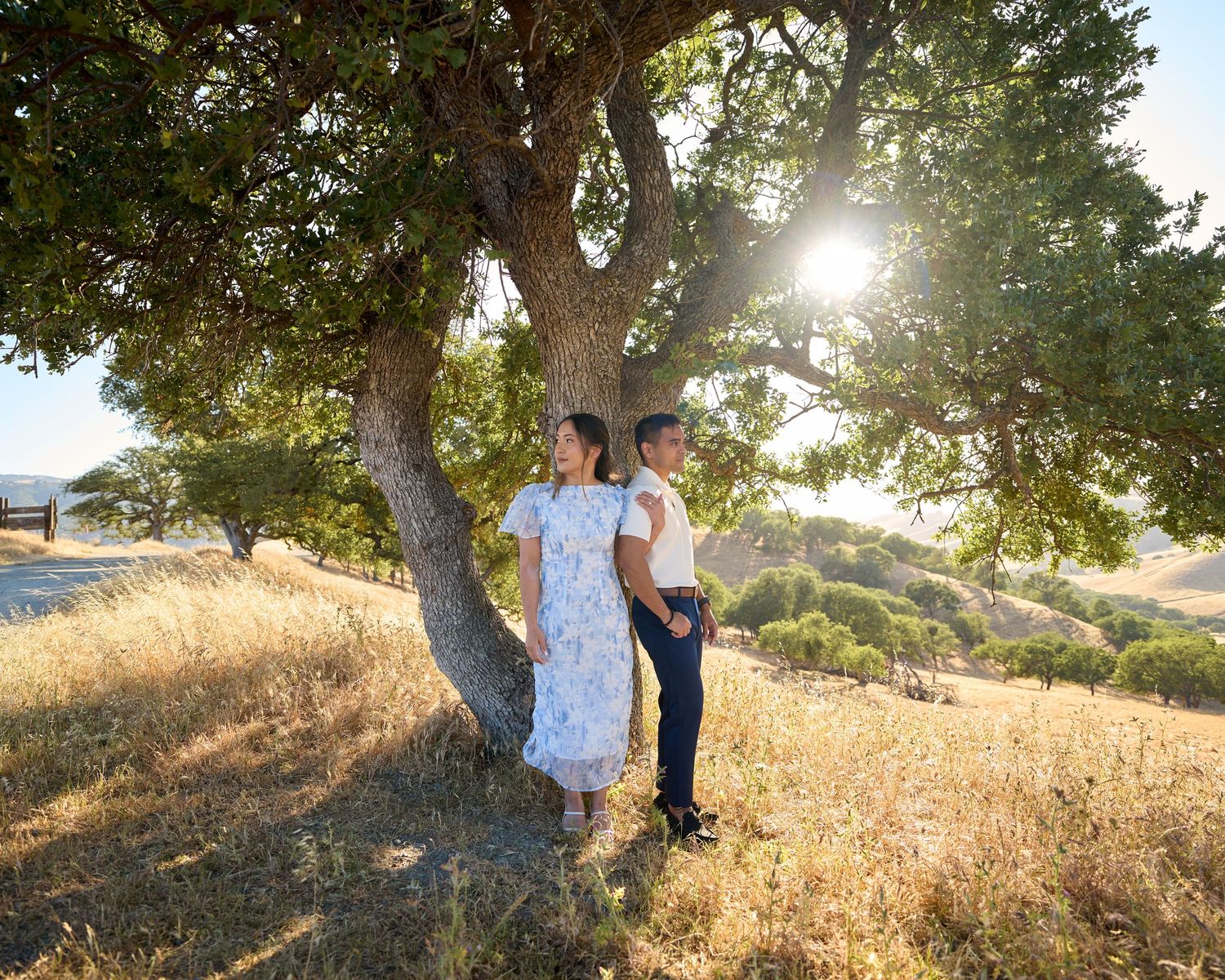 An engaged couple smiling opposite directions underneath a tree during sunset time.