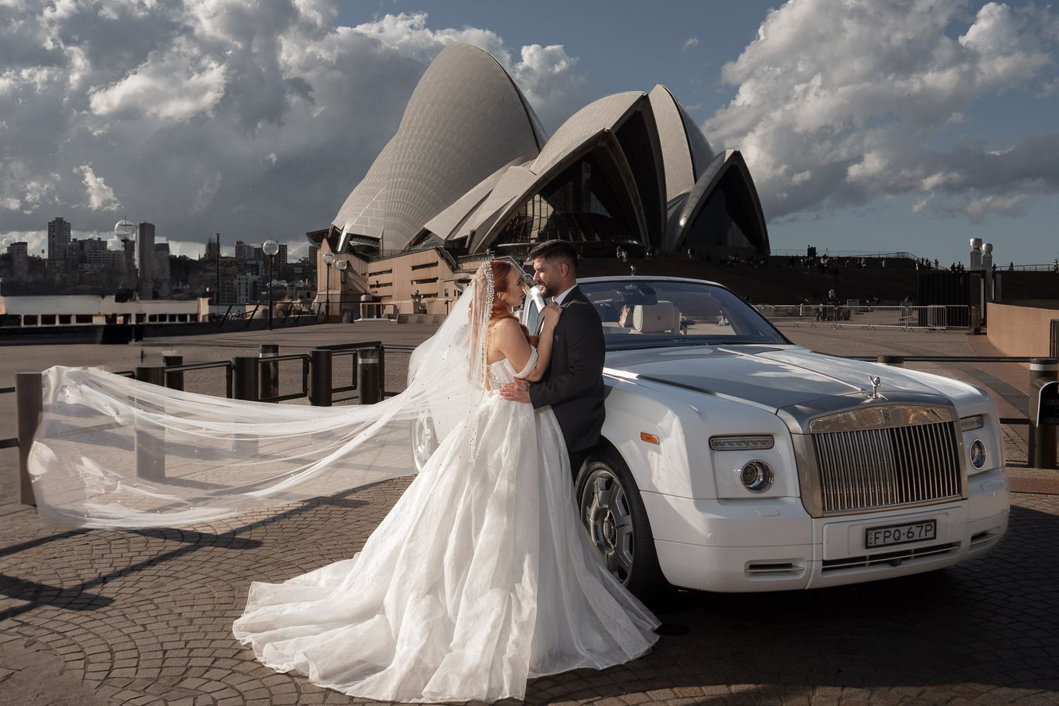A wedding couple poses with a white Rolls Royce in front of the Sydney Opera House.