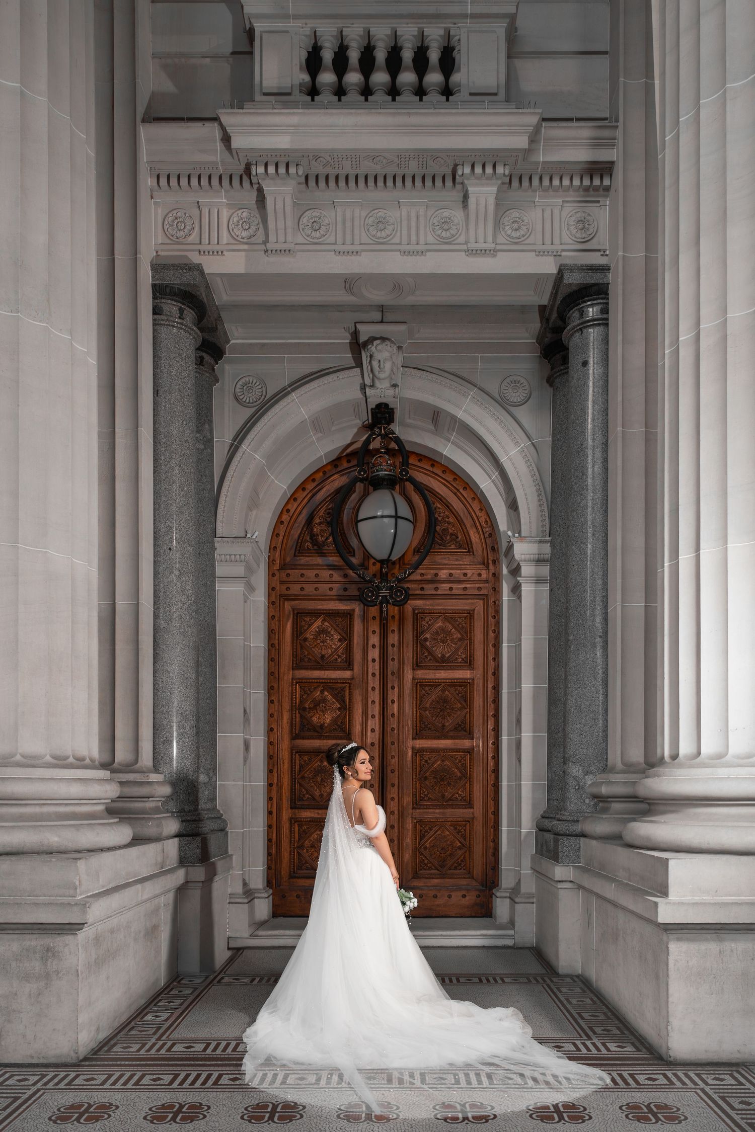 Bride in white gown and veil poses before ornate wooden arched door flanked by marble columns in elegant venue.