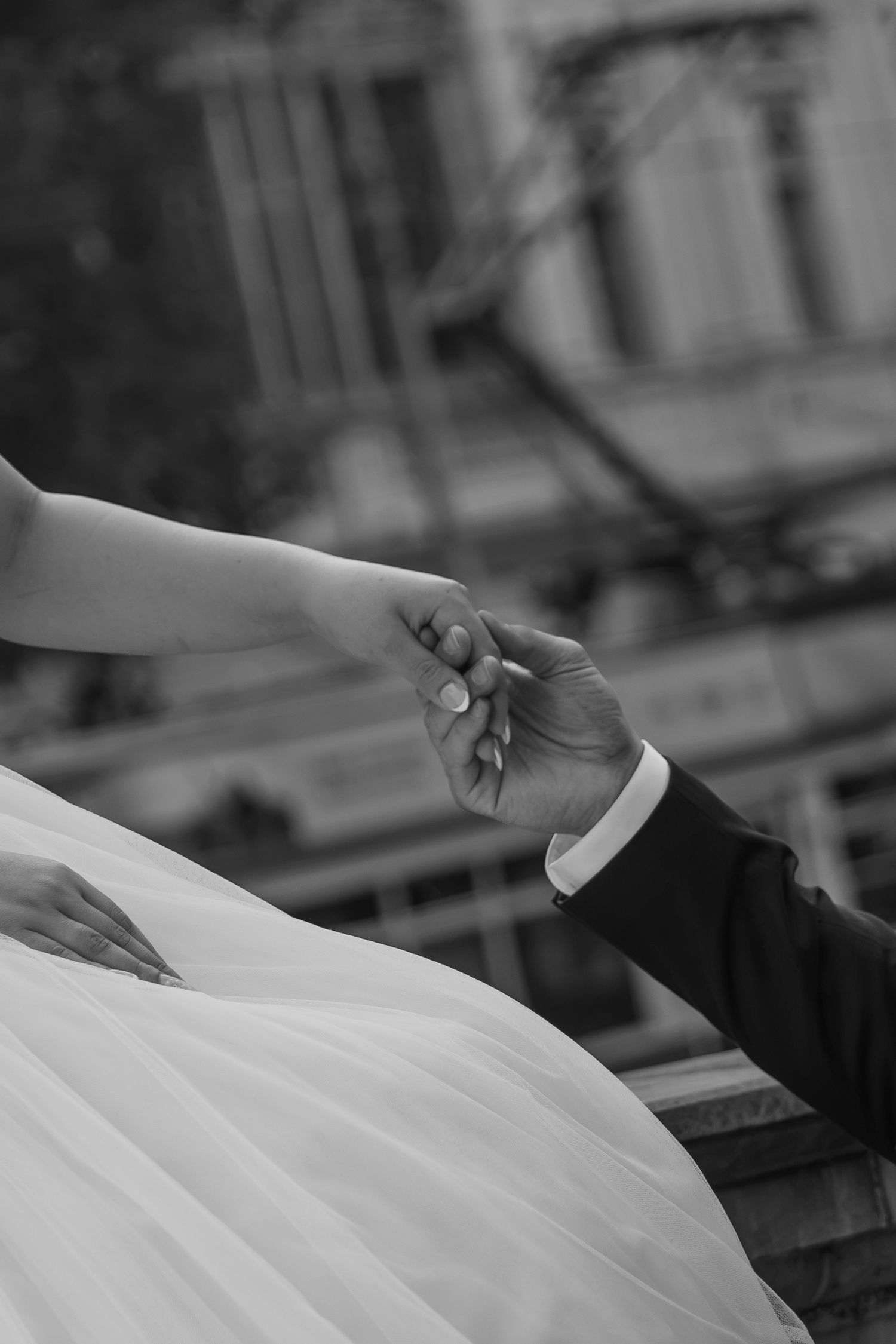 Black and white photo of bride and groom holding hands, wedding dress tulle visible, romantic outdoor ceremony moment.