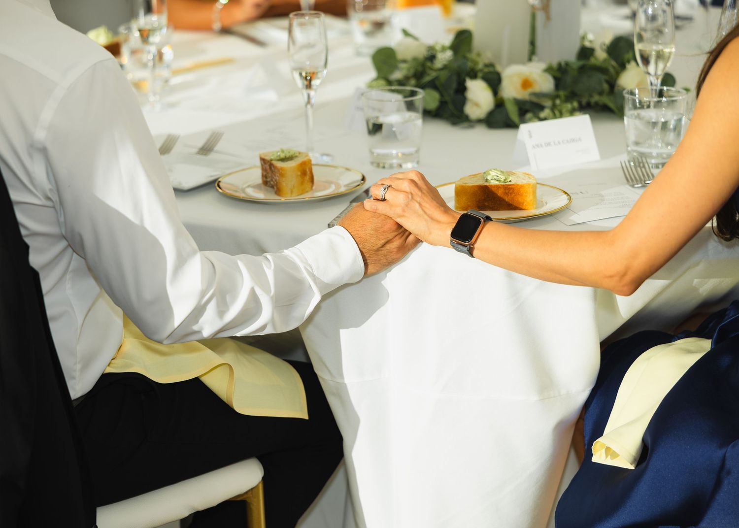 Two people holding hands across an elegant dining table set with plates and floral centerpieces at a formal event.