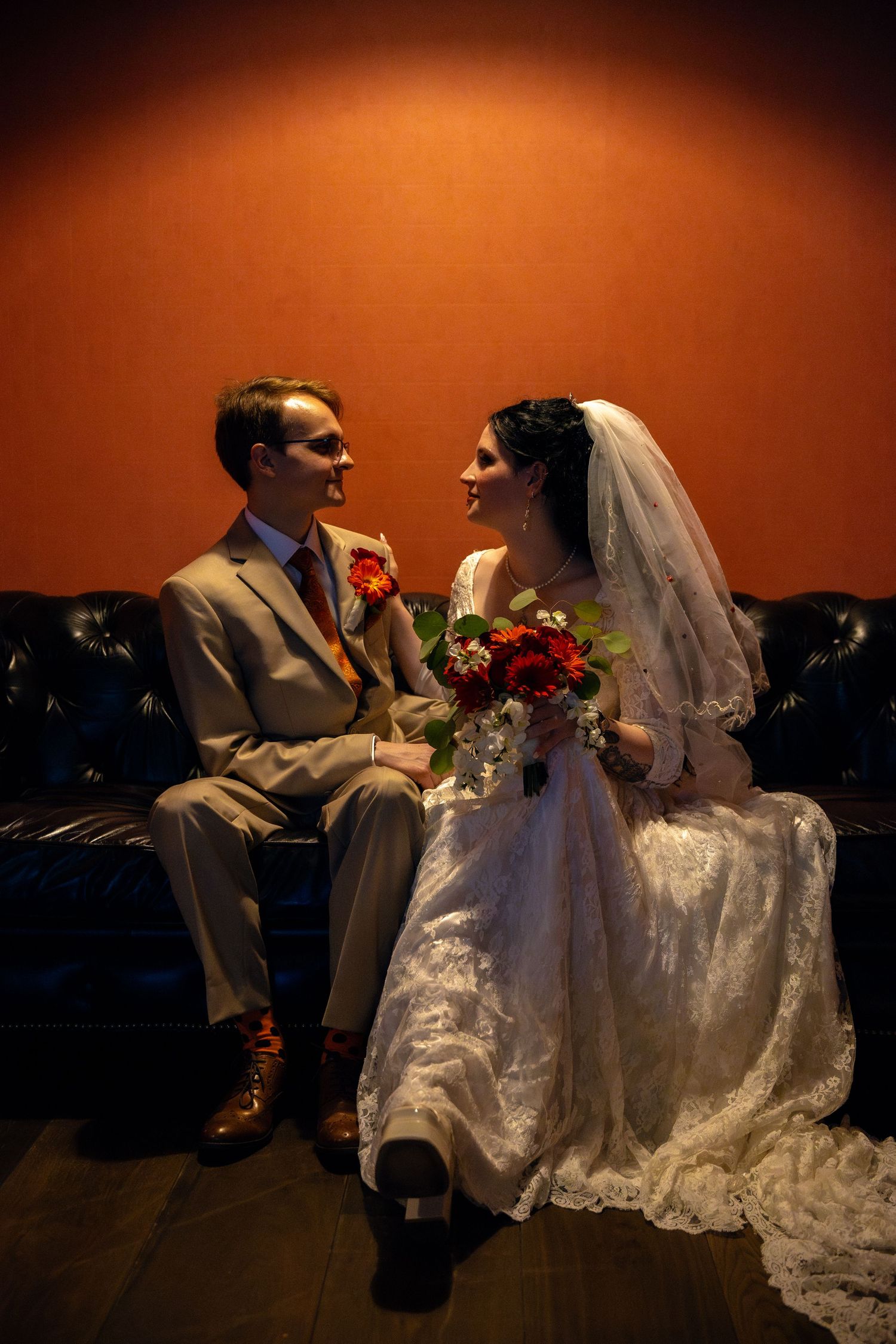 Wedding couple seated on dark leather couch against orange wall with red bouquet with dramatic lighting from above.