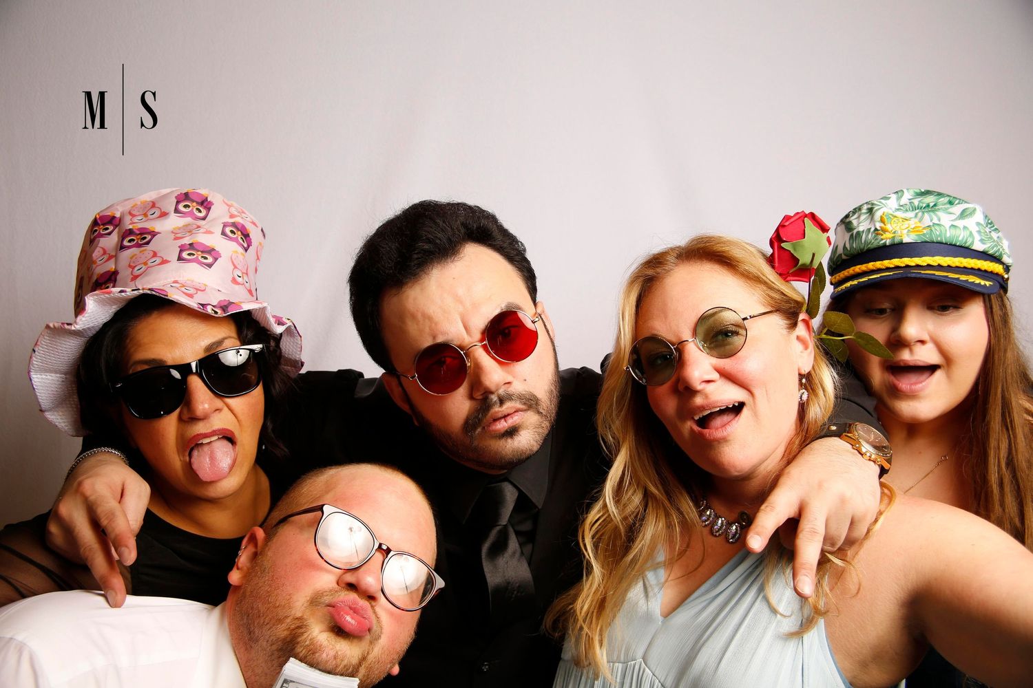 A group of friends wearing sunglasses and various hats pose together for a casual photo booth portrait.