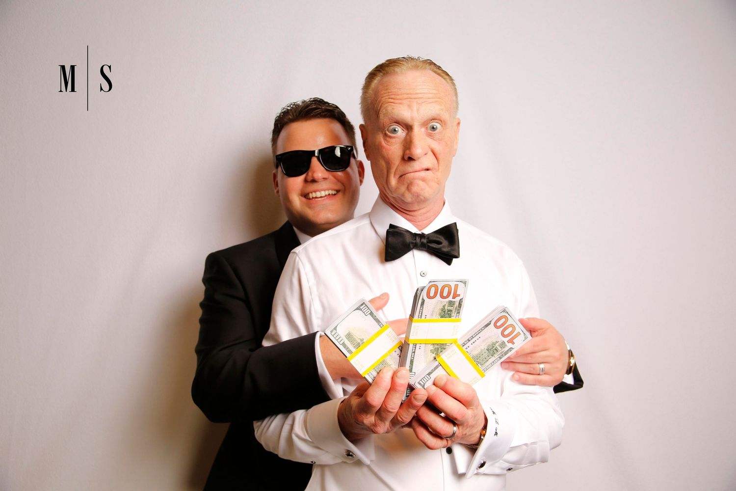 A couple in formal attire pose together with one holding stacks of money bills against a light gray background in a glam photo booth