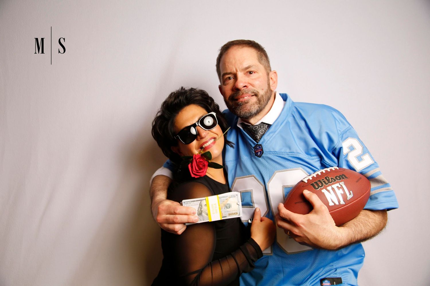A man wearing a Detroit Lions jersey poses in a photo booth with a woman wearing black sunglasses while holding a football and money