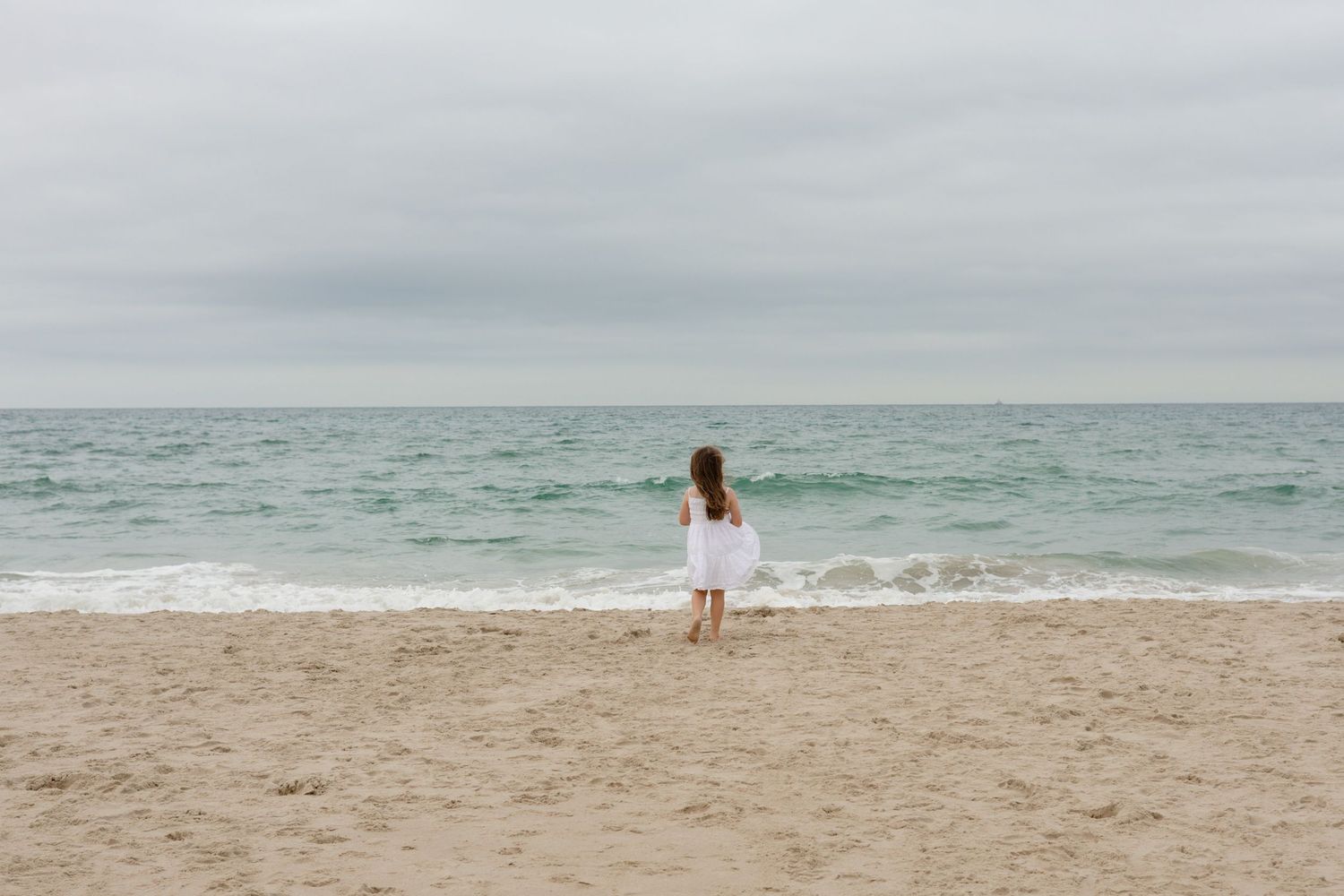 A young girl in a white dress stands alone on a sandy beach, facing the ocean under an overcast sky.
