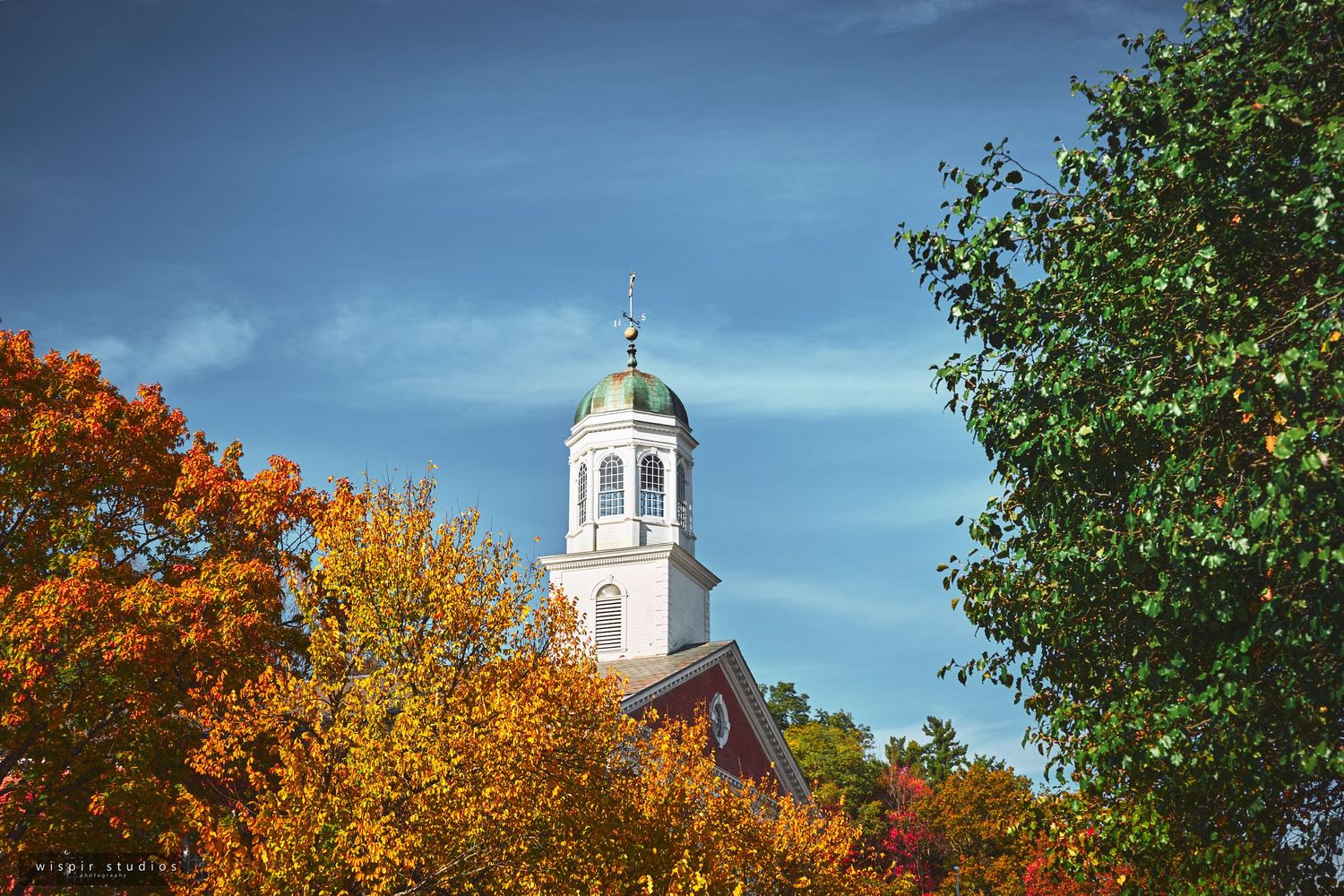 Historic white church steeple rises against bright blue sky with vibrant orange autumn foliage in foreground.