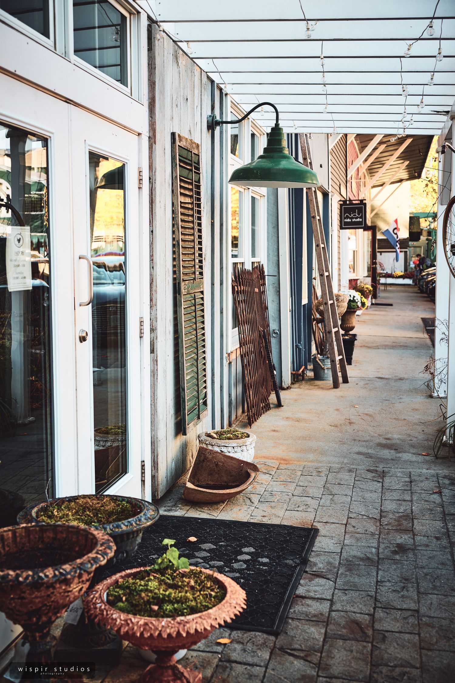 A rustic covered walkway with vintage green lamps and terracotta planters along a weathered storefront.