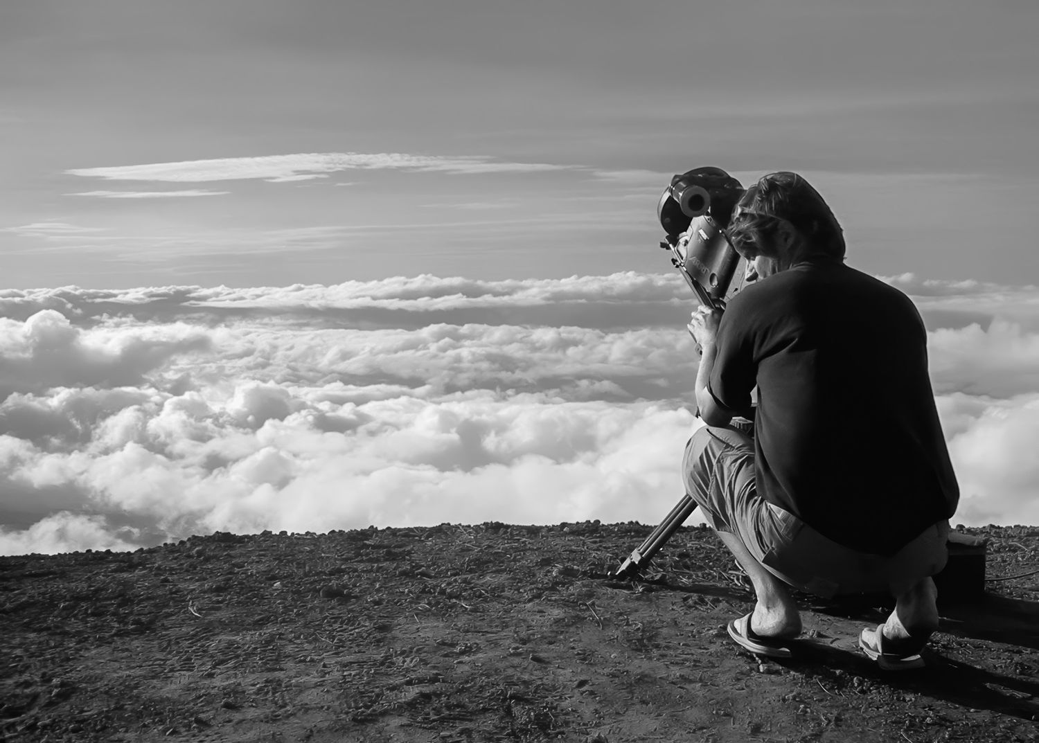 Silhouette of photographer crouched on mountain peak above clouds at sunset.