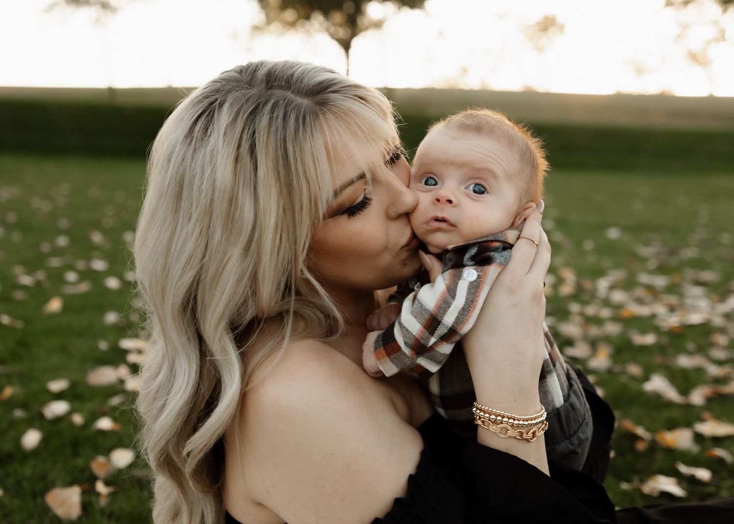 Close-up portrait of mother kissing baby on cheek in outdoor autumn setting.